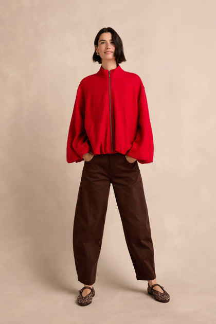 A woman stands with her hands in her pockets, wearing the Puffball Boiled Wool Jacket in red, paired with brown loose pants and leopard-print flats against a neutral background.