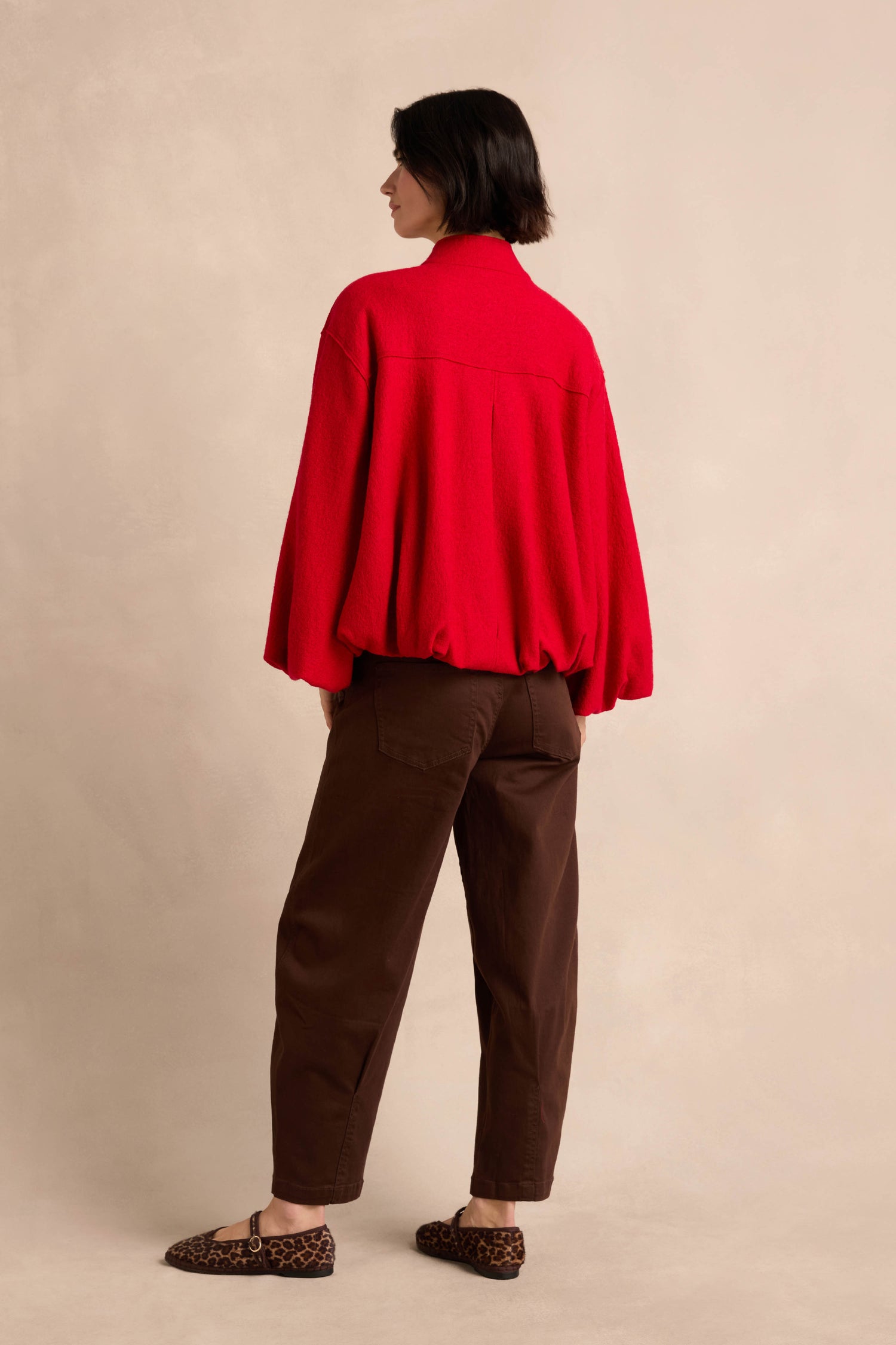 A person stands with their back to the camera, wearing the Puffball Boiled Wool Jacket in red, paired with brown pants and leopard print flats against a neutral background.