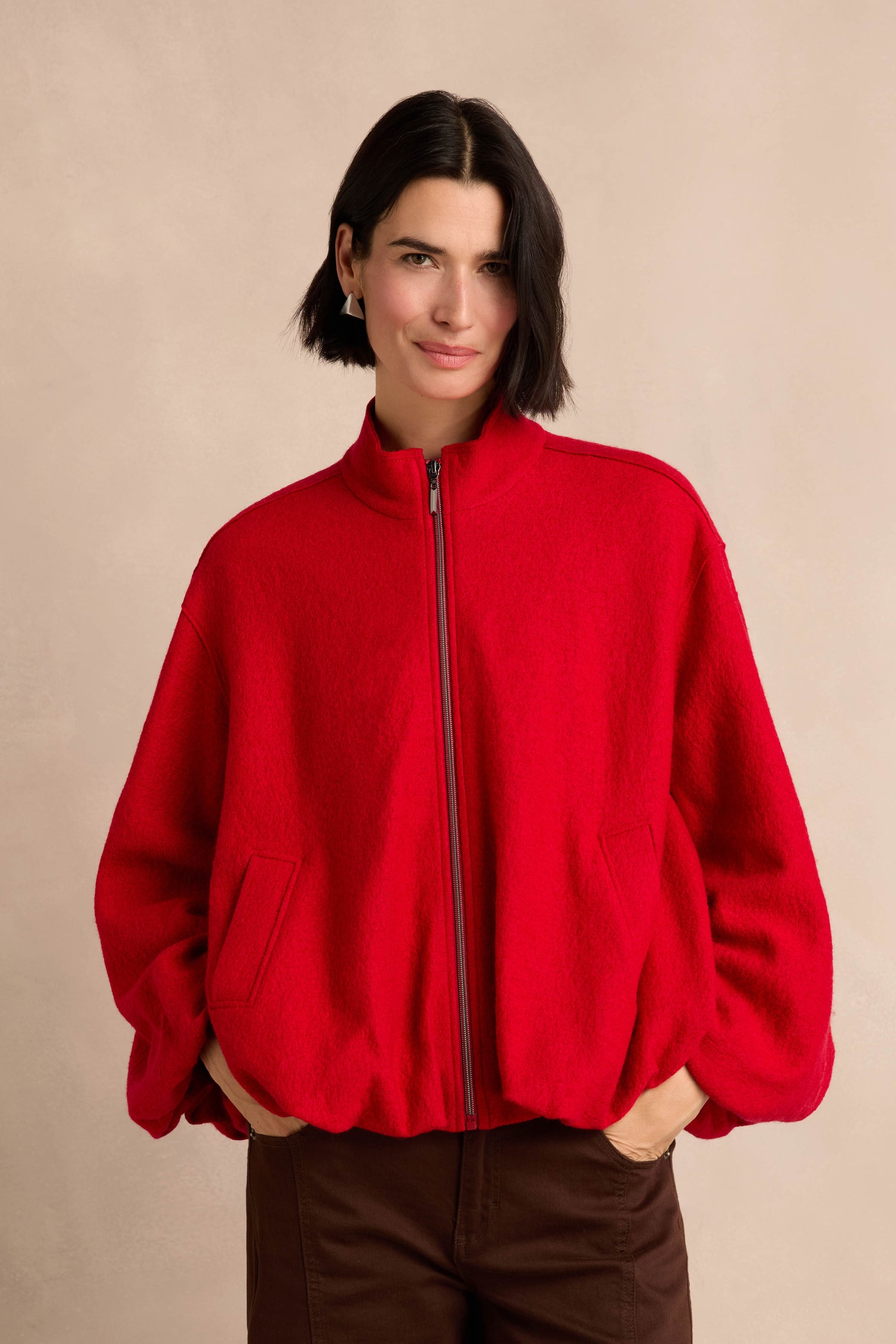 A person with short dark hair wears the Puffball Boiled Wool Jacket in bright red, paired with brown pants, standing against a plain beige background with hands in the jacket pockets.