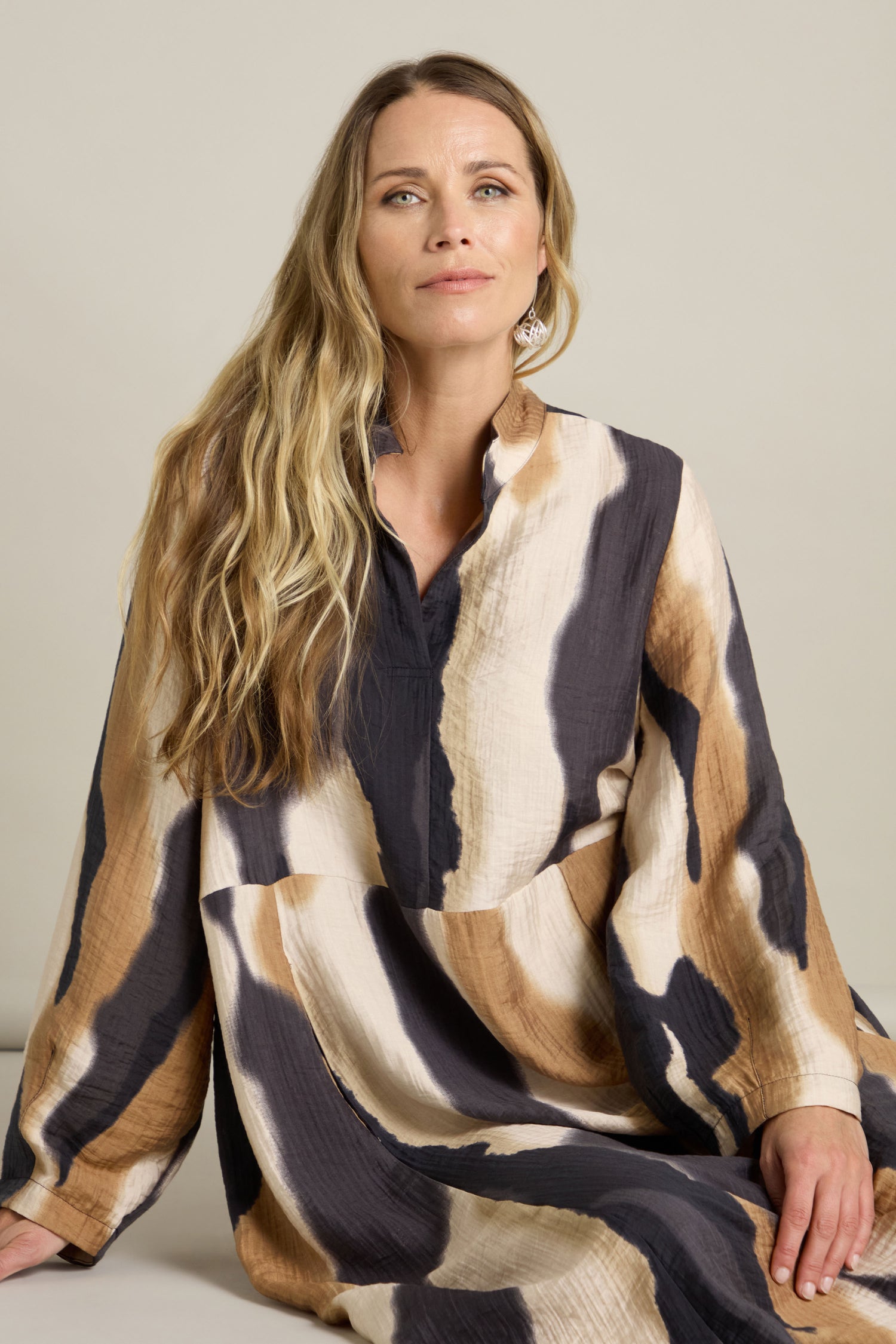 A woman is seated, wearing a Crinkle Viscose Print Dress featuring a stand open collar and an abstract pattern in black, brown, white, and beige, looking straight ahead.