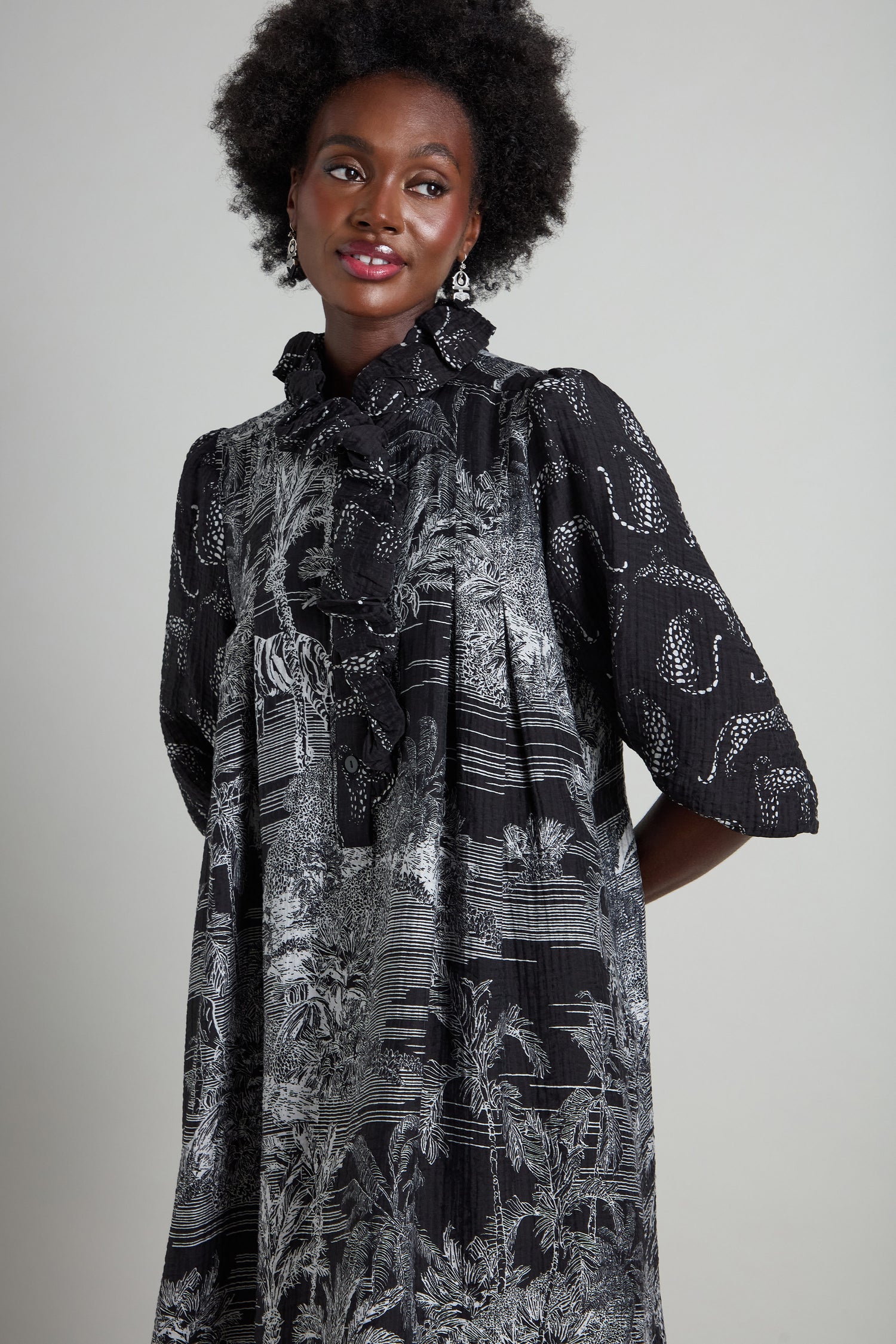 The individual is dressed in a Frill Neck Viscose Dress showcasing a jungle-themed black and white pattern, accented with elegant ruffles. This A-line silhouette garment highlights intricate designs as they stand against a plain backdrop, gazing slightly to the side.