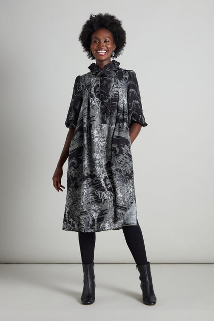 A smiling person stands against a plain background, highlighting the Frill Neck Viscose Dress with its jungle-inspired print and A-line silhouette. They pair it with black leggings and matching boots, which perfectly enhance the striking patterned design.