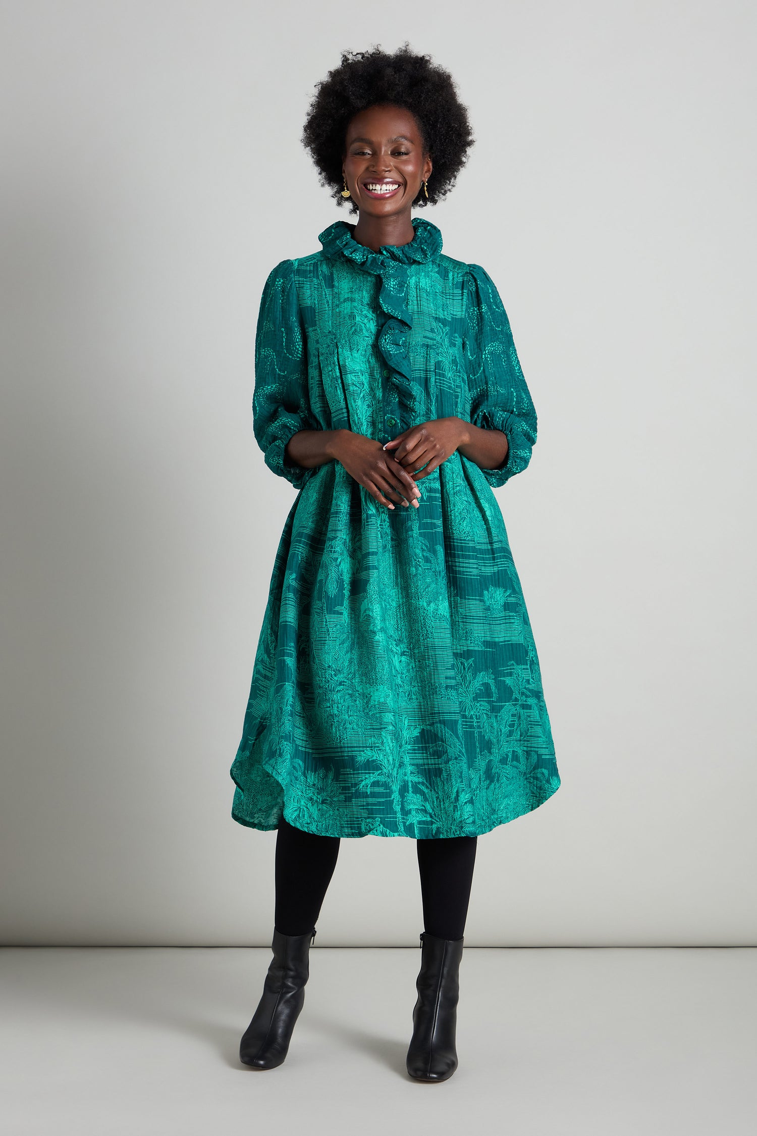 A smiling person stands against a plain background, wearing the Frill Neck Viscose Dress in a green pattern with an A-line silhouette, complemented by black leggings and boots.