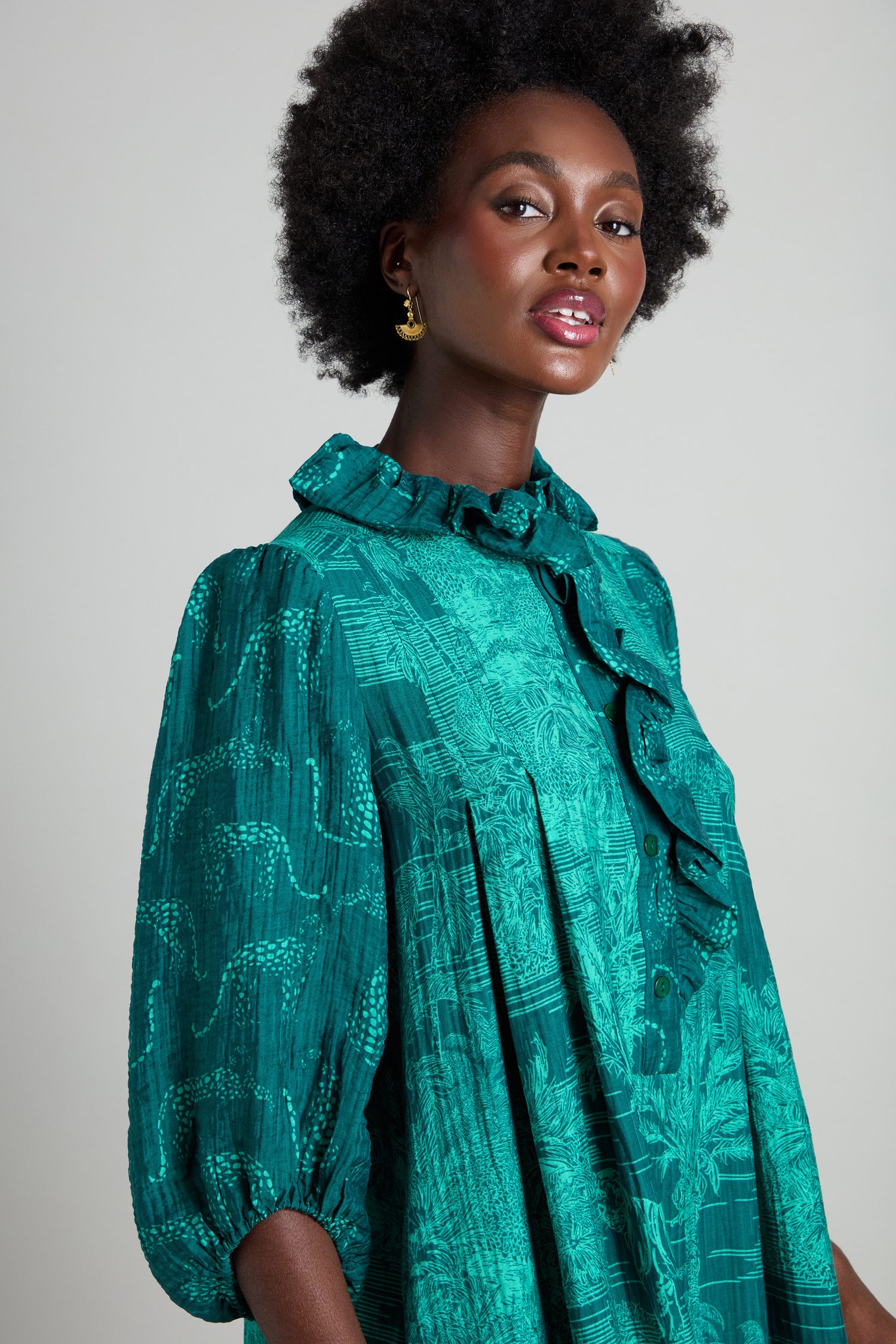 Against a neutral background, the person is wearing a Frill Neck Viscose Dress with a jungle-inspired print. The teal pattern showcases an A-line silhouette, enhanced by ruffled collar and sleeves, seamlessly combining natural motifs with chic design.