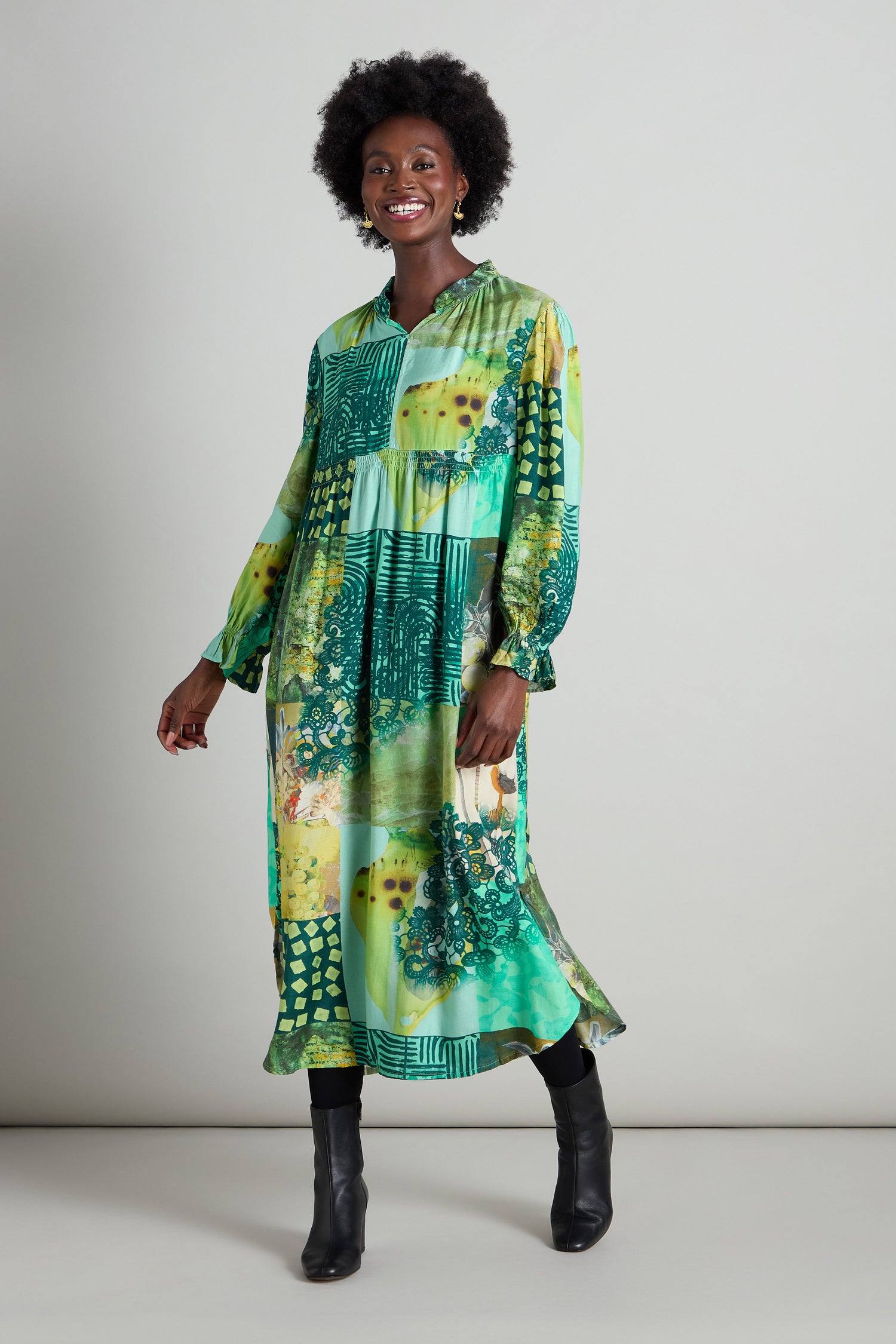 A person stands smiling in a Patchwork Print Viscose Dress, featuring vibrant green hues with floral and geometric patterns, beautifully complemented by an empire waistline, against a neutral background.