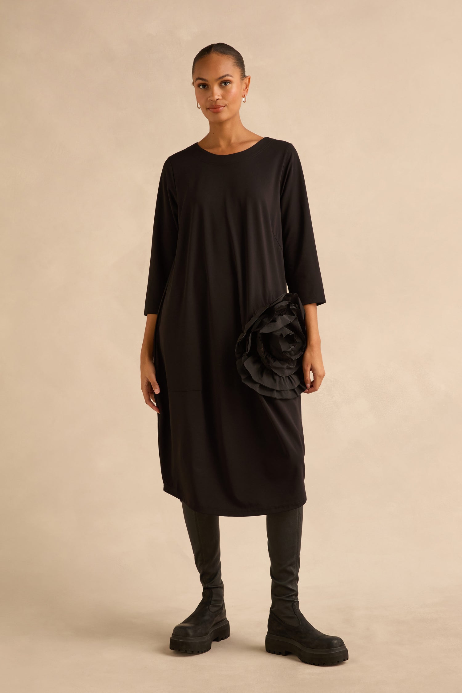 A person wearing a black Taffeta Flower Detail Jersey Dress in a cocoon silhouette with a large fabric flower at the hip, black leggings, and black lug-sole boots.