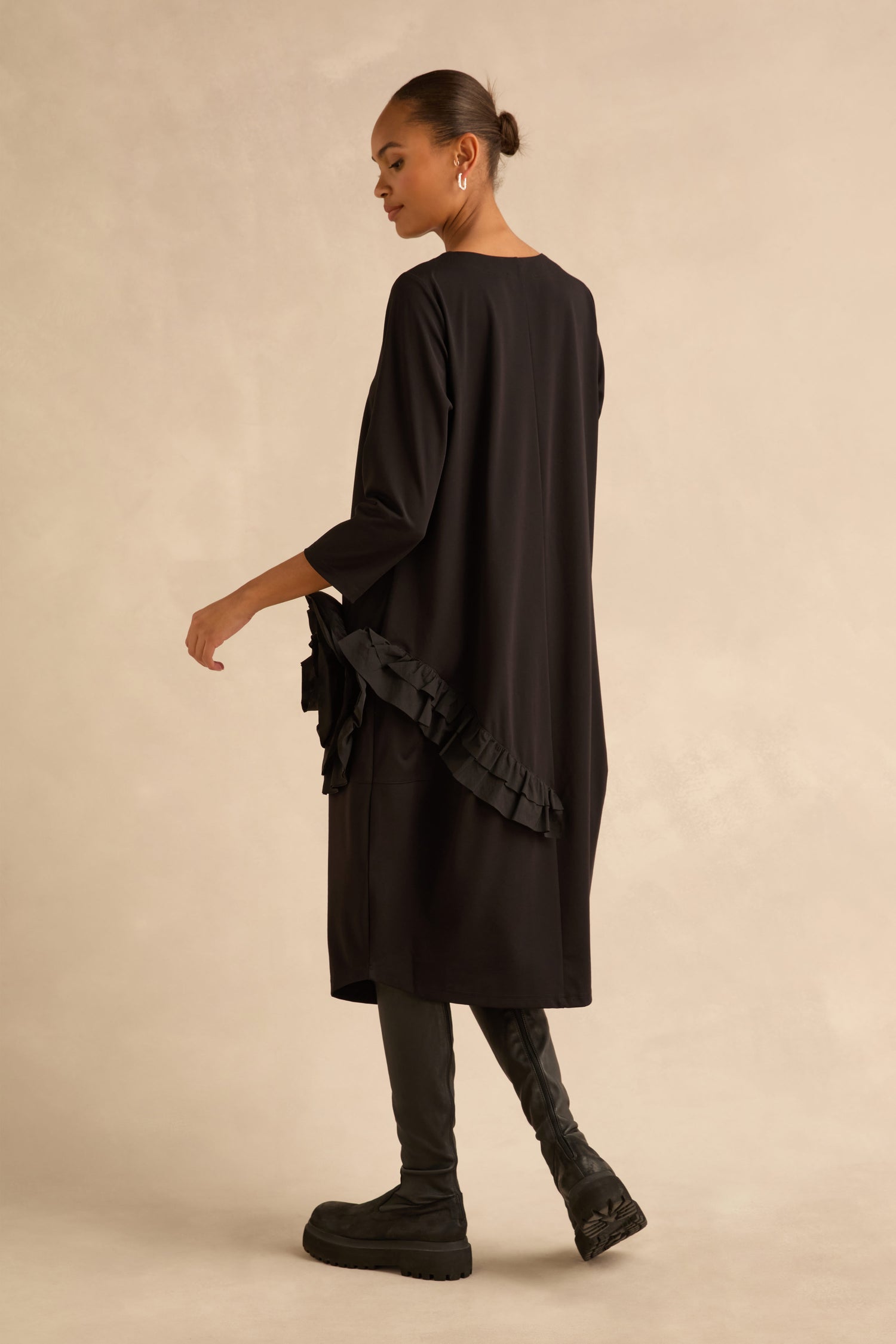 A person wearing the black Taffeta Flower Detail Jersey Dress in a cocoon silhouette with a ruffled hip accent, paired with black boots.