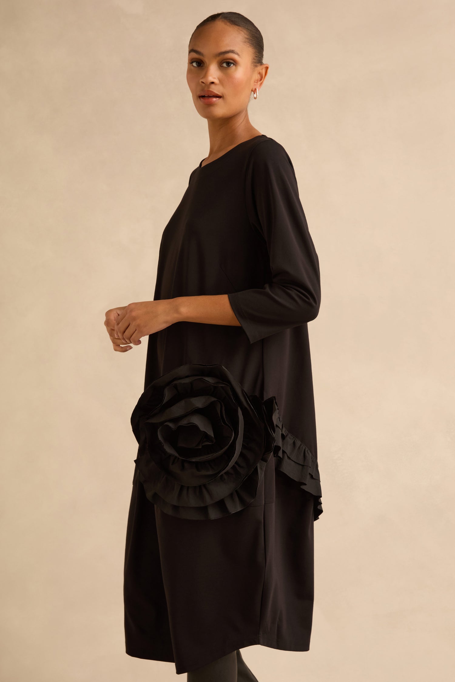 A model wears the Taffeta Flower Detail Jersey Dress, a black jersey dress in a cocoon silhouette with three-quarter sleeves and a large, dimensional fabric flower detail at the hip.