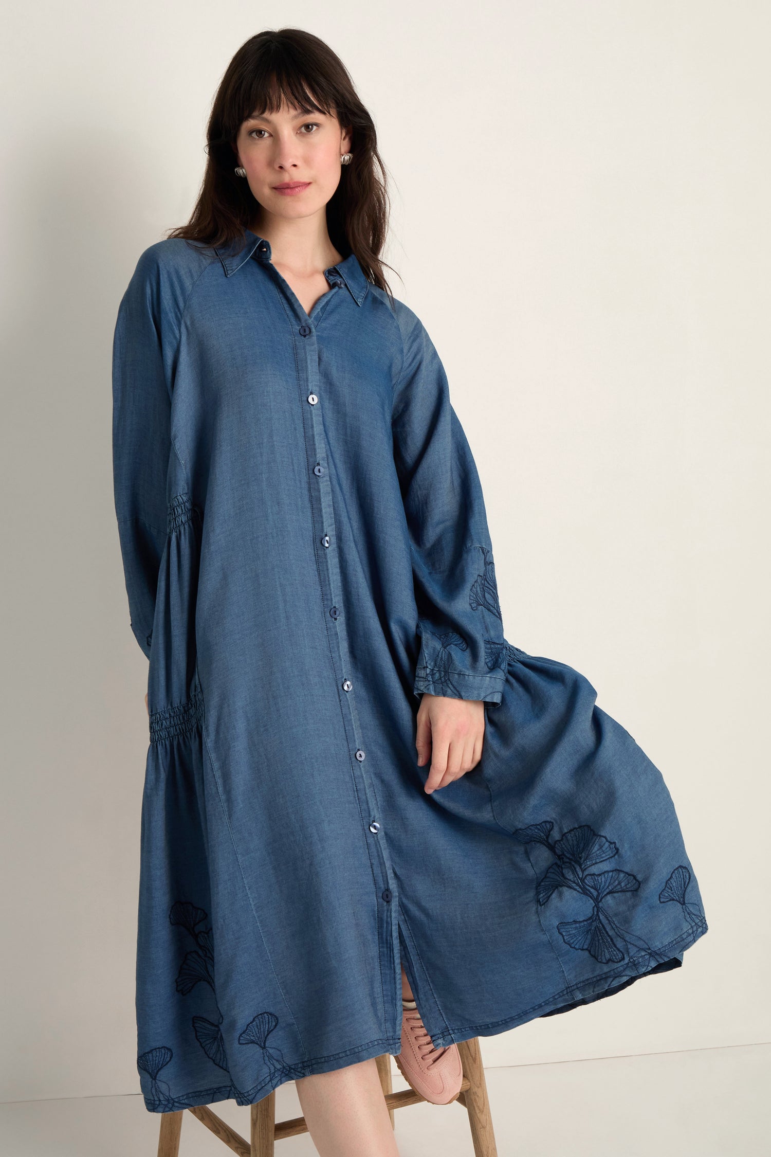 A woman wears the Ginkgo Embroidered Denim Dress, a relaxed, long-sleeved midi made from soft lyocell denim with subtle embroidery, standing against a plain light background.