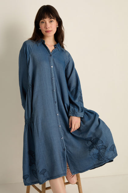 A woman wears the Ginkgo Embroidered Denim Dress, a relaxed, long-sleeved midi made from soft lyocell denim with subtle embroidery, standing against a plain light background.