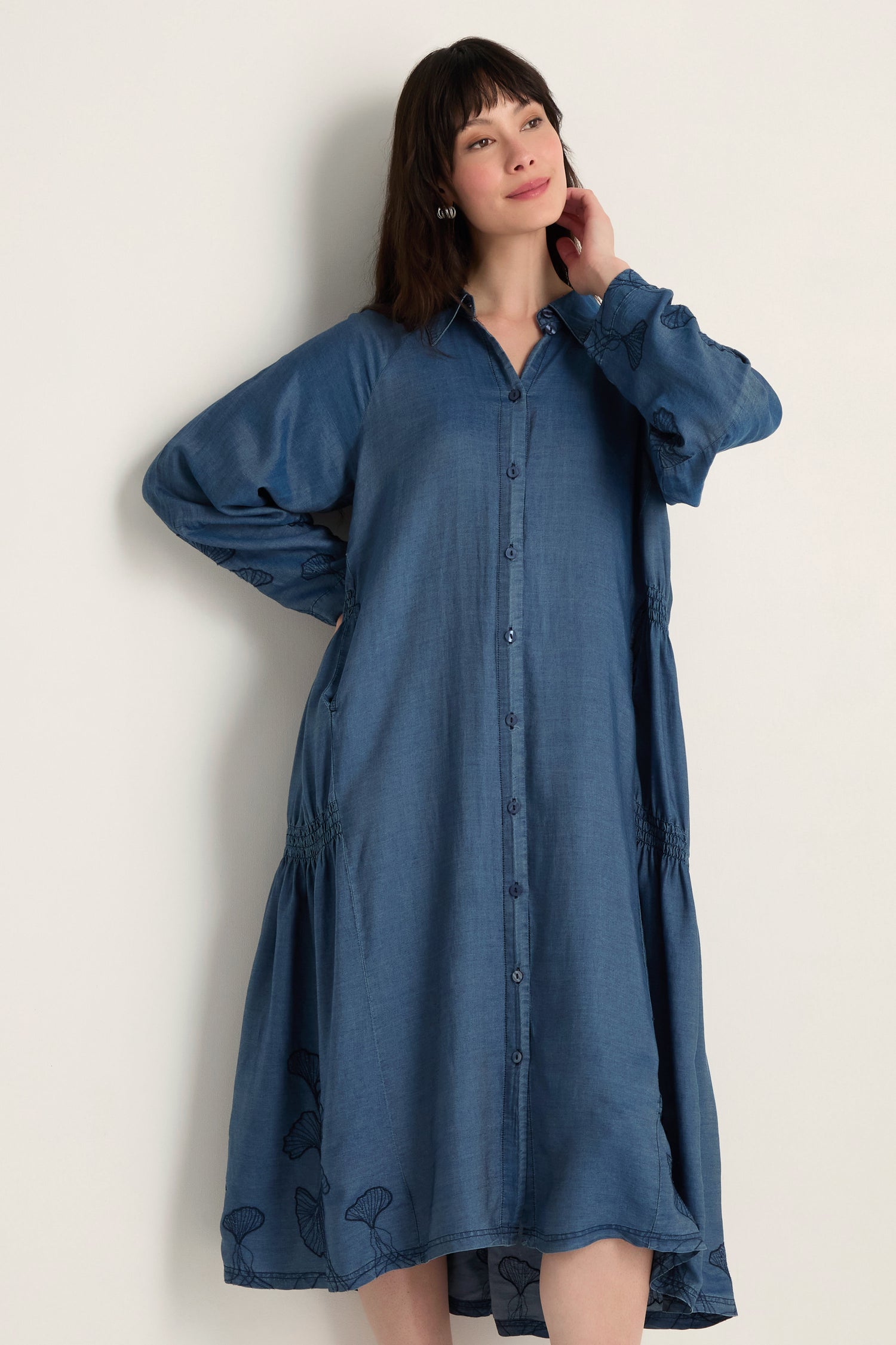 A woman poses against a plain background, wearing the Ginkgo Embroidered Denim Dress—a loose-fitting, long-sleeved, midi dress crafted from lyocell denim.
