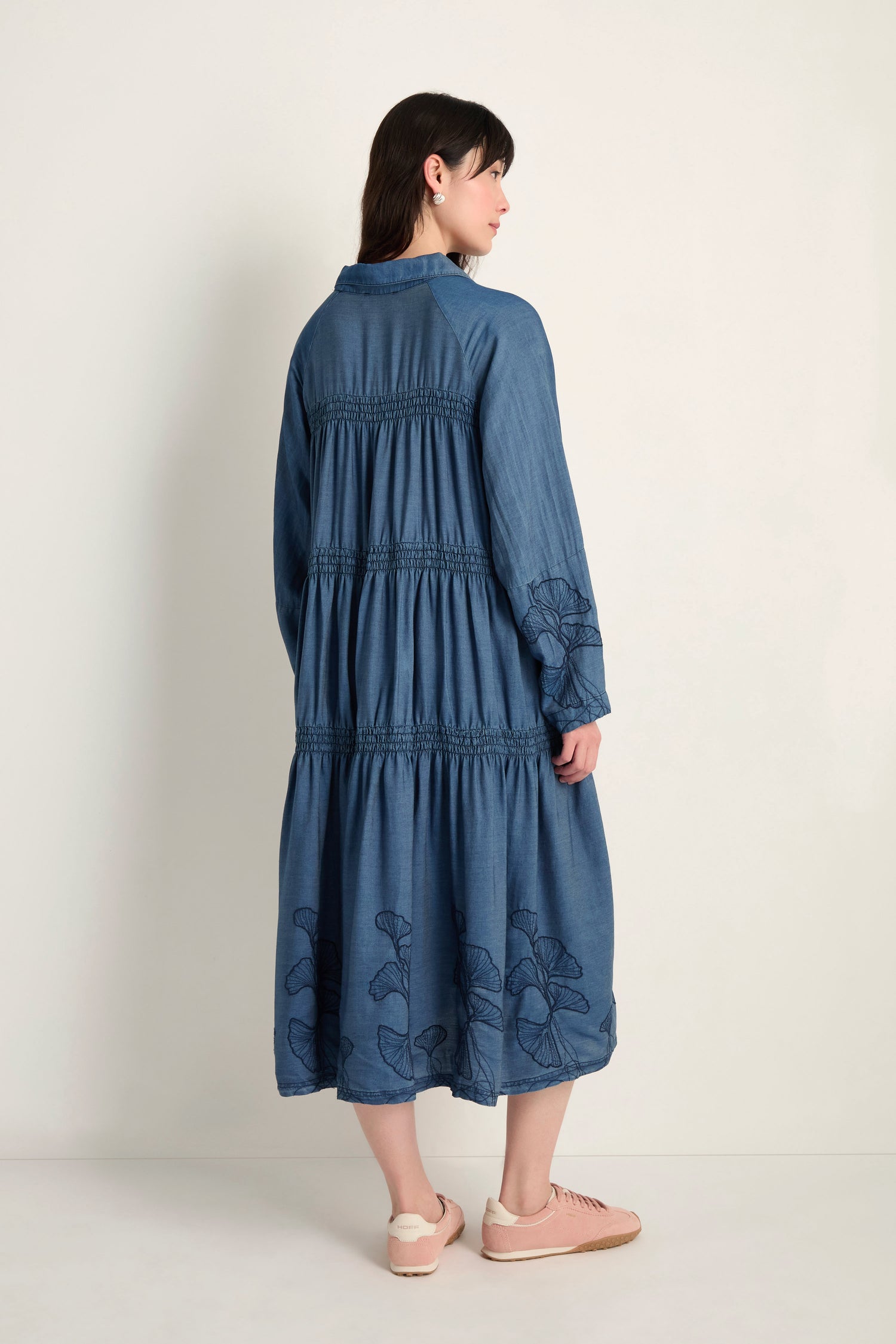 A woman stands facing away, wearing the Ginkgo Embroidered Denim Dress in midi length with gathered details and floral embroidery, paired with light pink sneakers.
