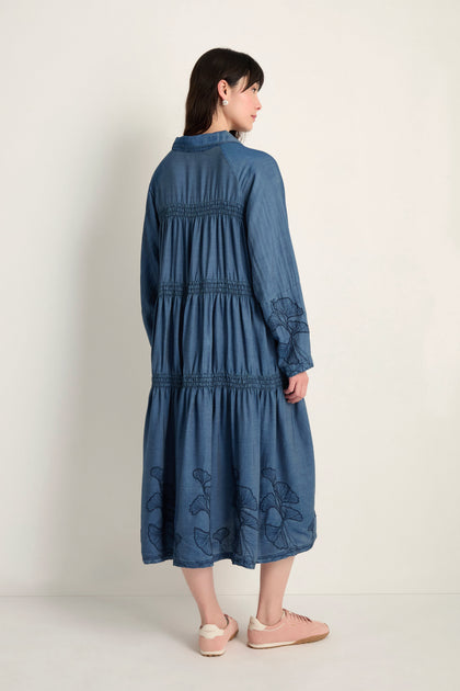 A woman stands facing away, wearing the Ginkgo Embroidered Denim Dress in midi length with gathered details and floral embroidery, paired with light pink sneakers.