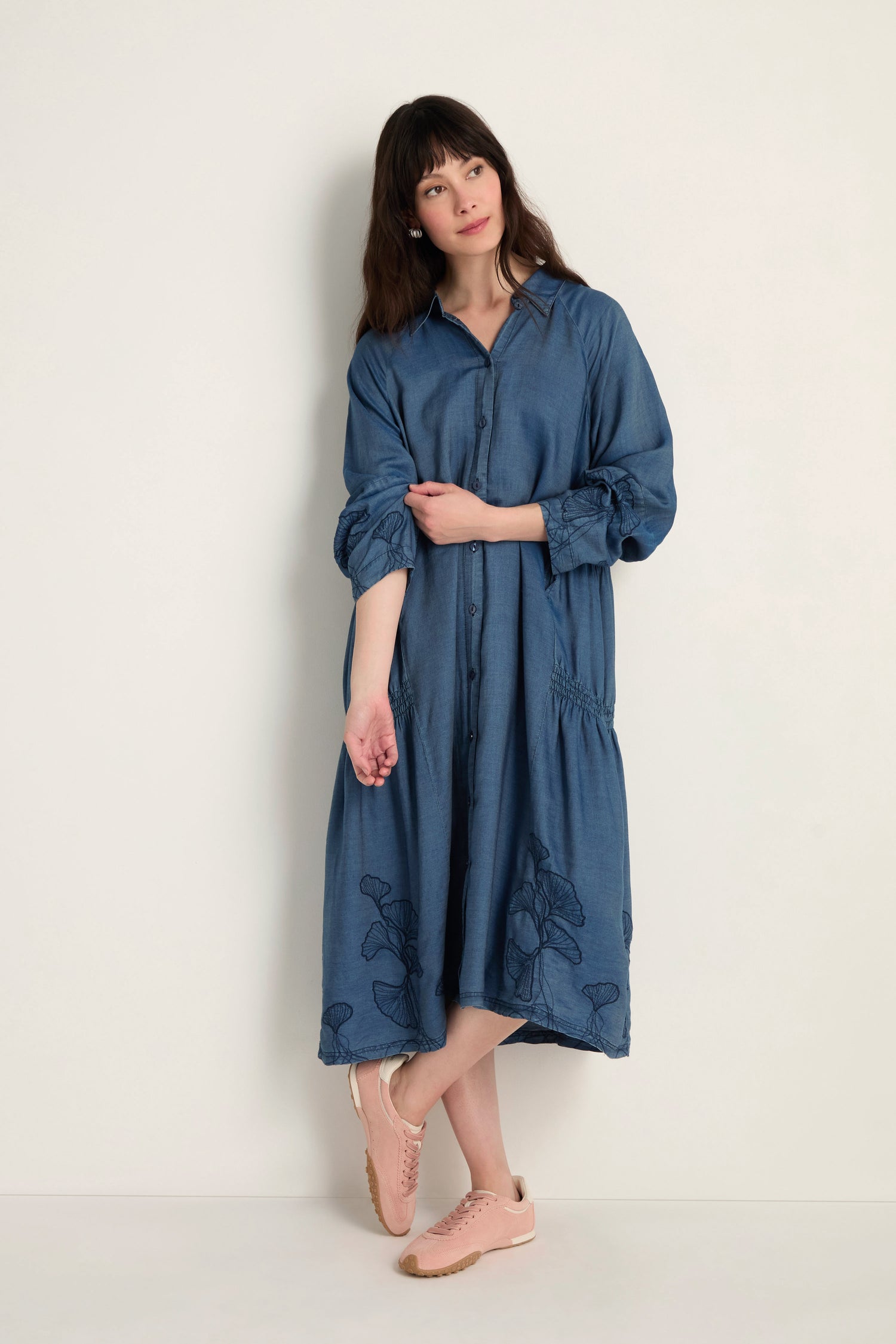 A woman stands by a light wall wearing the loose-fitting midi Ginkgo Embroidered Denim Dress with button front and floral accents, paired with pink sneakers.
