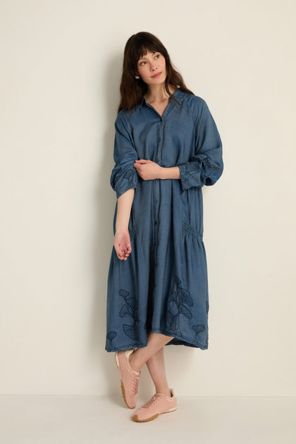 A woman stands by a light wall wearing the loose-fitting midi Ginkgo Embroidered Denim Dress with button front and floral accents, paired with pink sneakers.