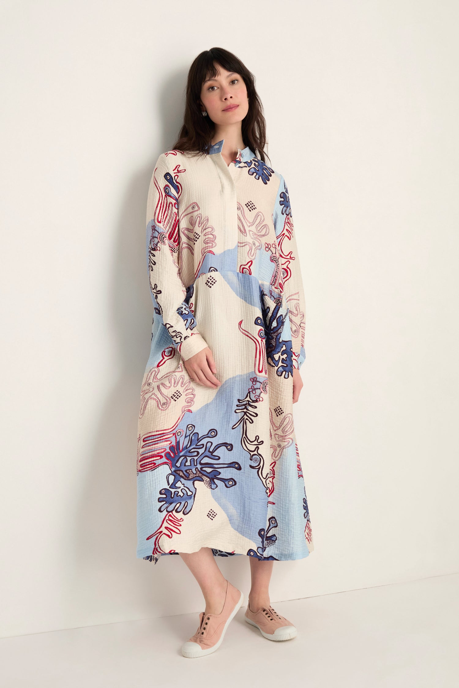 A woman stands against a white wall wearing the Abstract Botanical Crinkle Dress—a long-sleeved, collared dress with a relaxed silhouette in blue, cream, and red—paired with beige sneakers.
