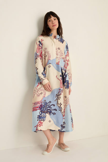 A woman stands against a white wall wearing the Abstract Botanical Crinkle Dress—a long-sleeved, collared dress with a relaxed silhouette in blue, cream, and red—paired with beige sneakers.