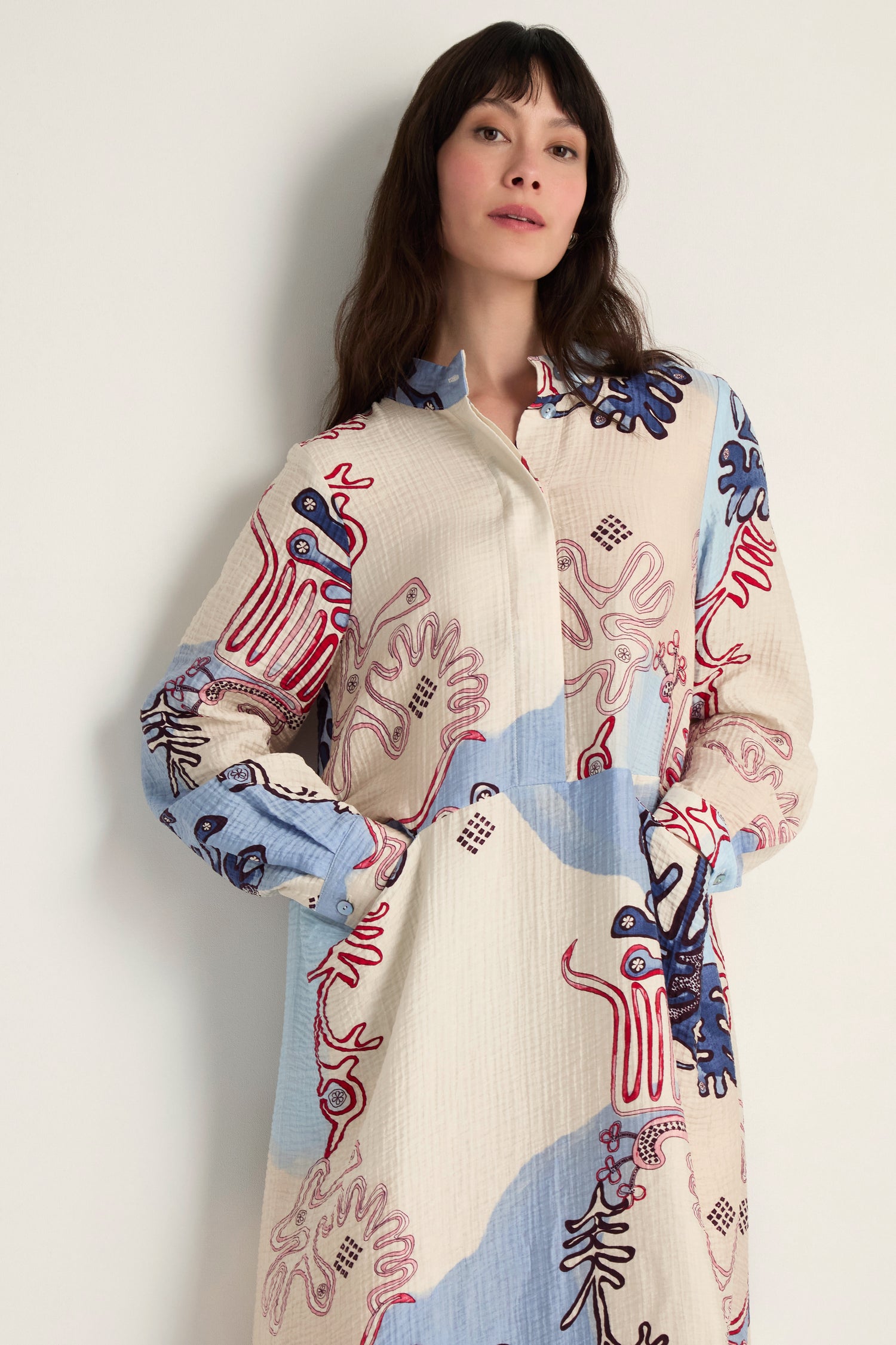 A woman stands against a light wall wearing the Abstract Botanical Crinkle Dress, a long-sleeved, button-up piece with a relaxed silhouette and abstract red, blue, and beige patterns.
