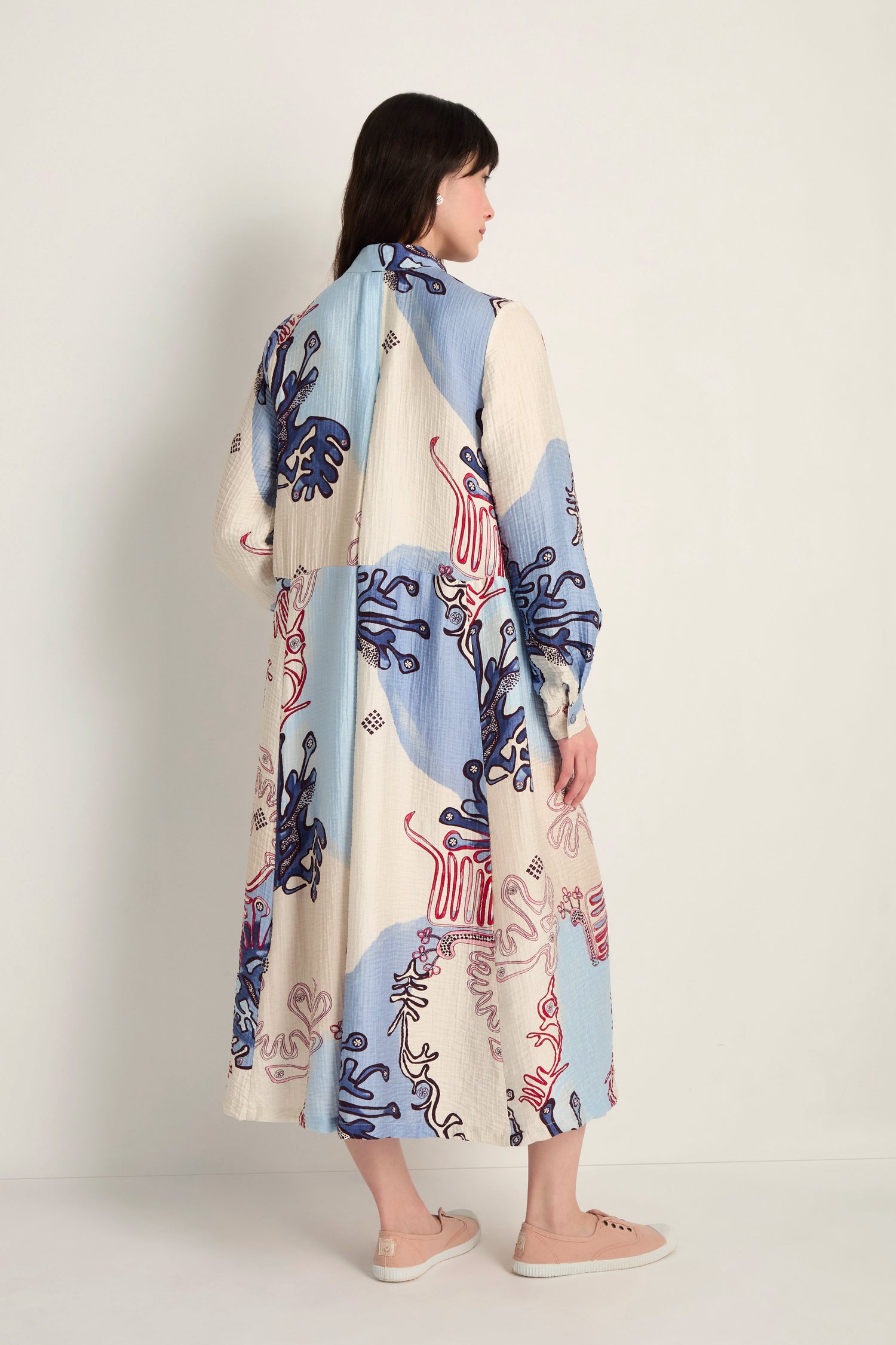 A woman faces away, barefoot on a light floor against a neutral wall, wearing the Abstract Botanical Crinkle Dress—blue, cream, and red with an abstract botanical print, long sleeves, ankle-length hem, and relaxed silhouette.
