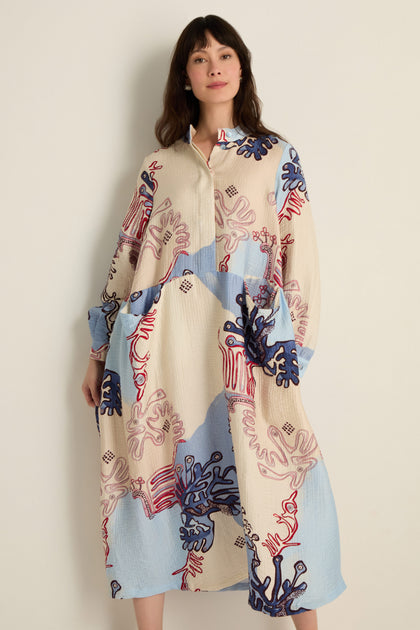 A woman wears the Abstract Botanical Crinkle Dress, featuring a relaxed silhouette, collared neckline, loose long sleeves, and abstract red, blue, and beige coral-inspired patterns.