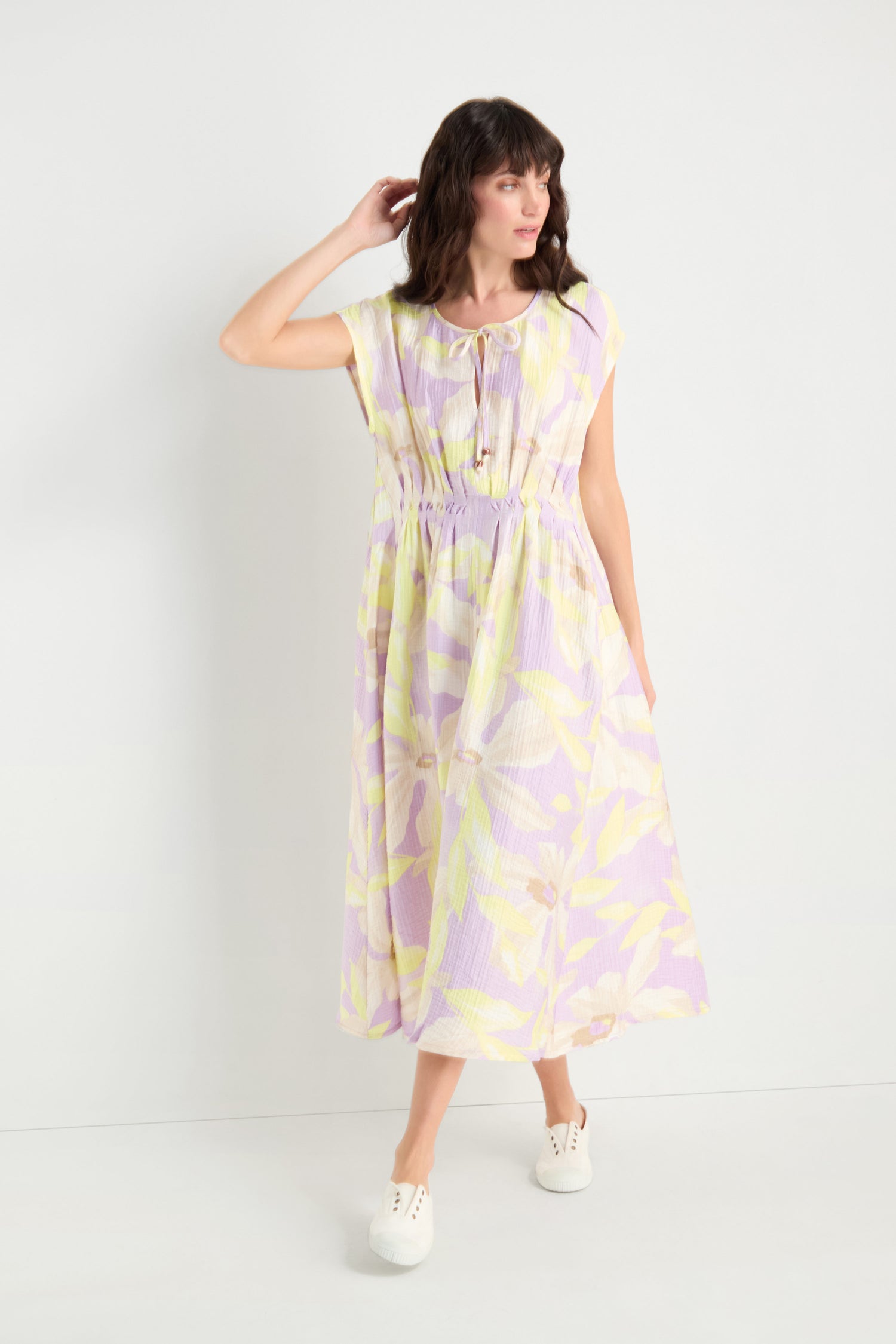 A woman stands against a plain white background wearing the Pleated Waist Print Dress, a pastel floral viscose blend midi with short sleeves, paired with white sneakers.