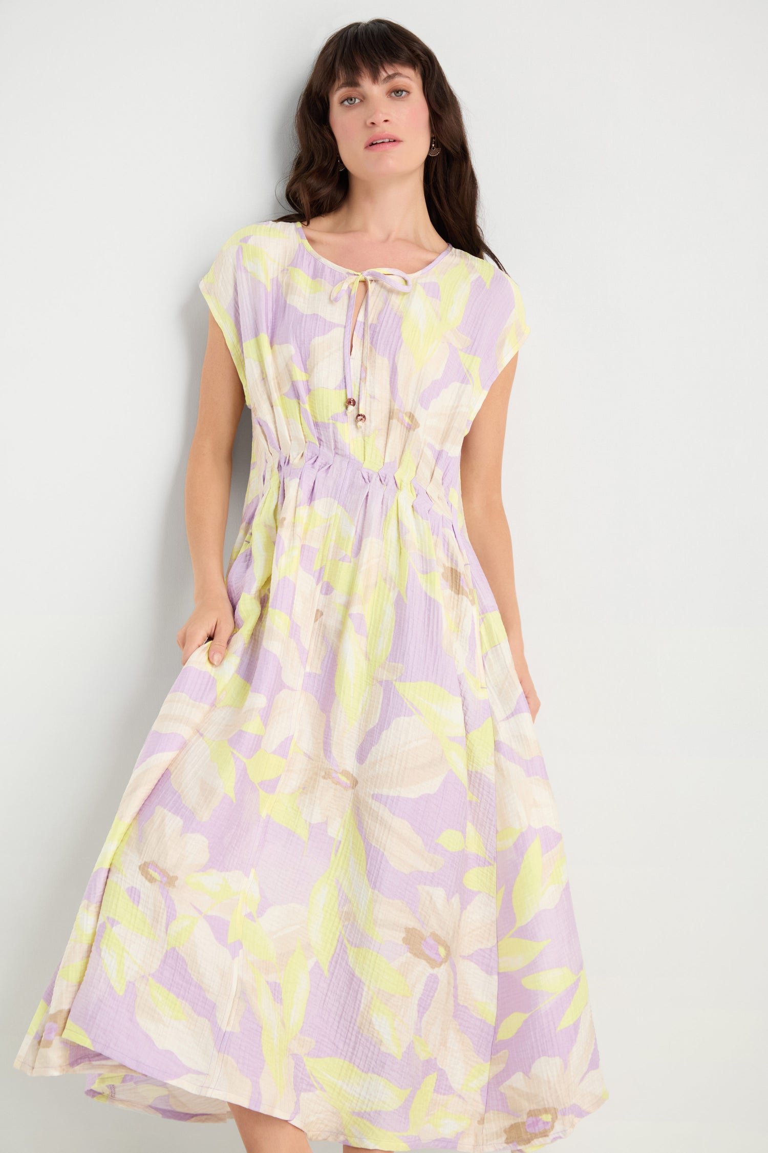 A woman models the Pleated Waist Print Dress, featuring a pastel floral pattern, viscose blend fabric, short sleeves, pleated waist, and tie neckline against a plain light-colored background.