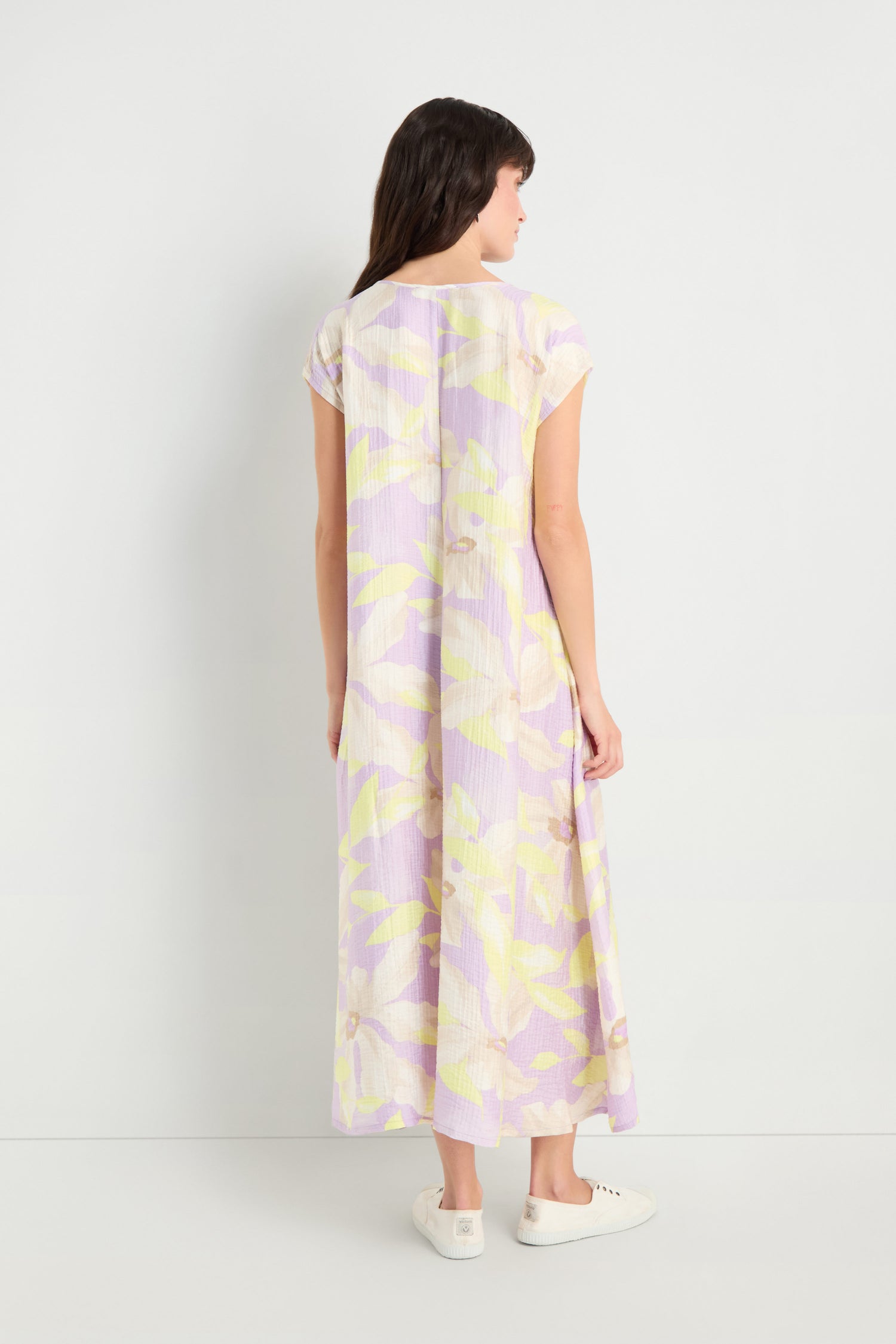 A woman, seen from behind, wears the Pleated Waist Print Dress—a short-sleeve, ankle-length, pastel floral viscose blend—paired with white sneakers in front of a plain white wall.
