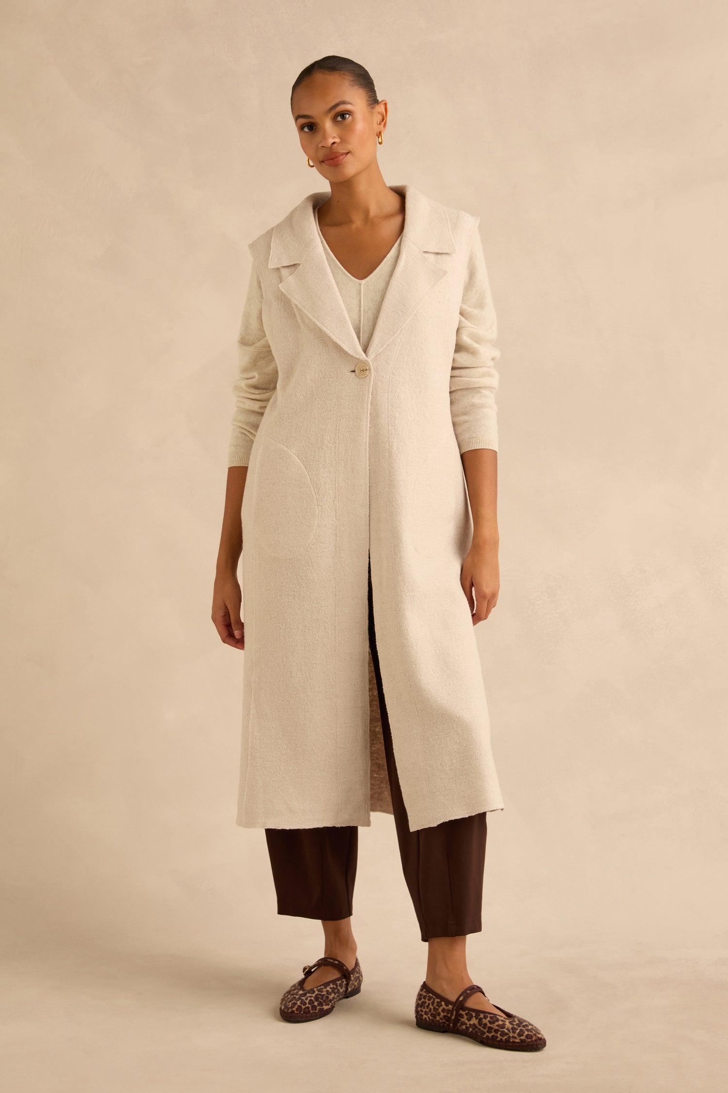 A person wearing a cream-colored wool blend Detachable Fur Long Gilet over a matching top and brown trousers, paired with leopard-print flats.