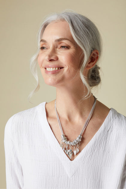 Mature woman with grey hair smiling gently, wearing a white top and a statement Silver Hearts Charm Necklace with adjustable length.