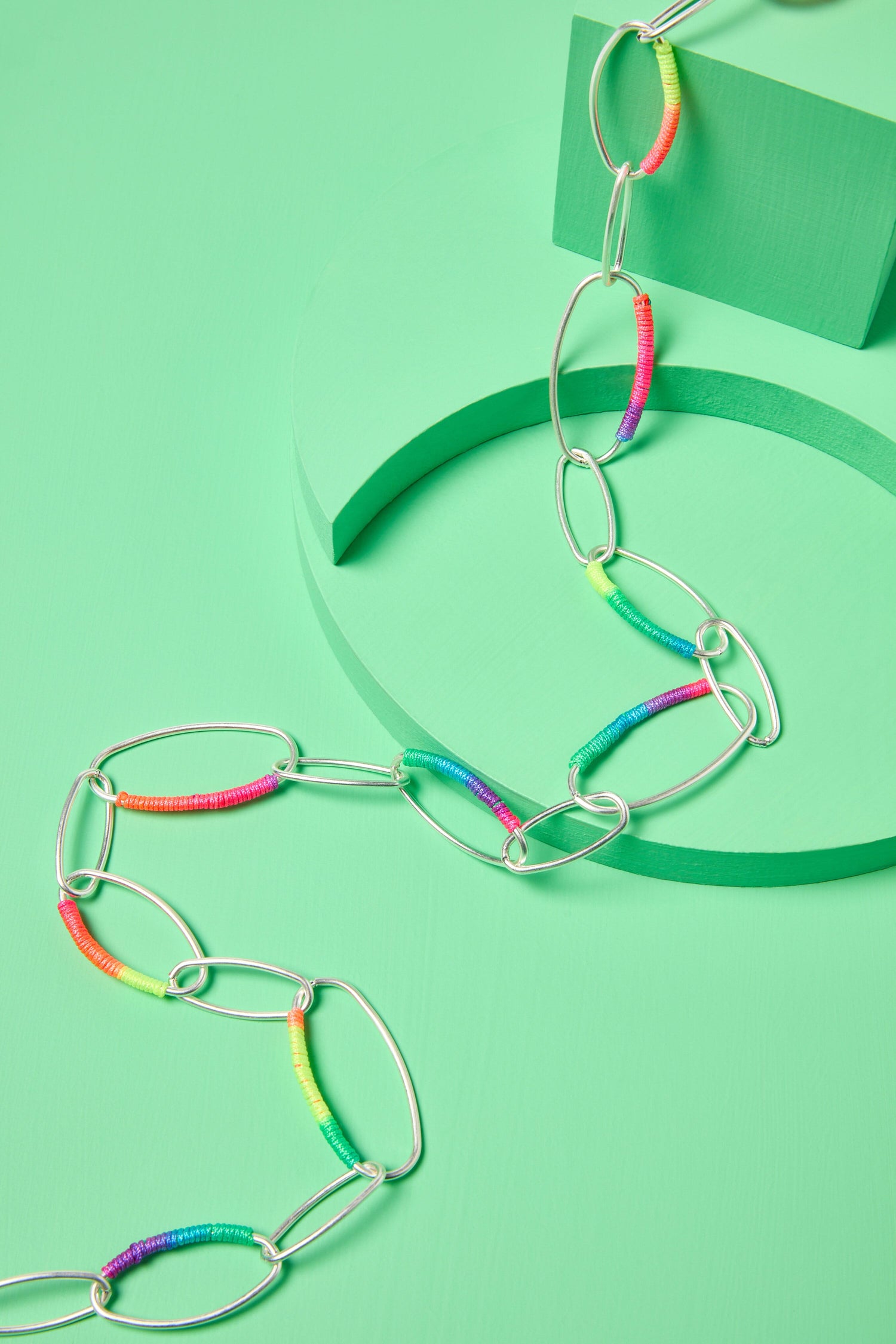 A Rainbow Stitch Silver Hoops Necklace on a pastel green background with geometric shapes.