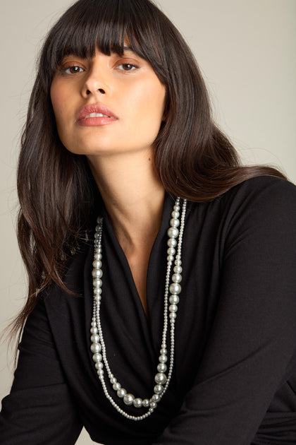 A woman with long dark hair wears the Graduated Silver Pearl Necklace layered over her black top, looking directly at the camera. The elegant design of the pearls adds a touch of sophistication to her classic look.