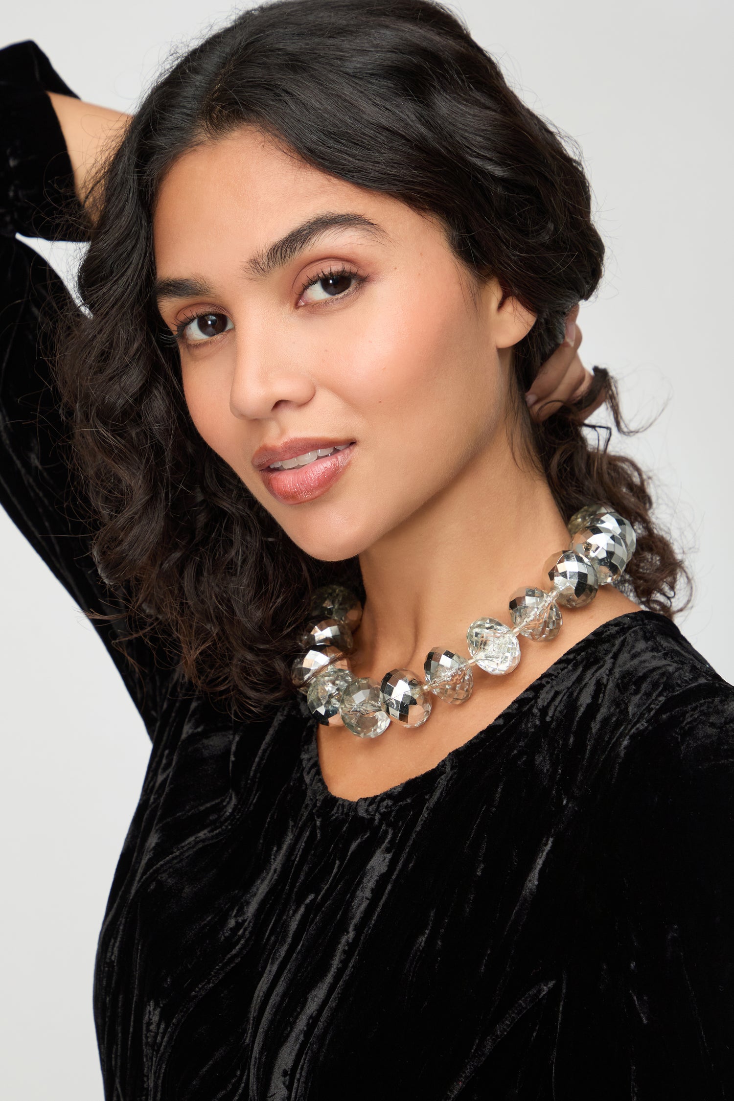 Woman with wavy dark hair wears a black velvet top and the Silver Disco Necklace, a chunky silver statement piece, while looking at the camera with a slight smile.