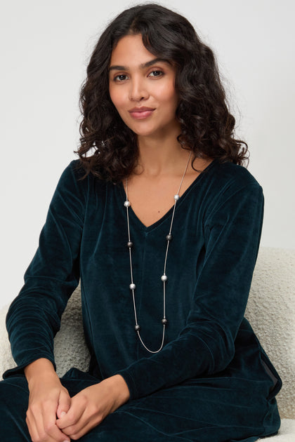 A woman with curly hair sits on a light chair, wearing a dark teal velvet top and the Sparkle Spheres Necklace, featuring silver and black beads, looking at the camera with a neutral expression.