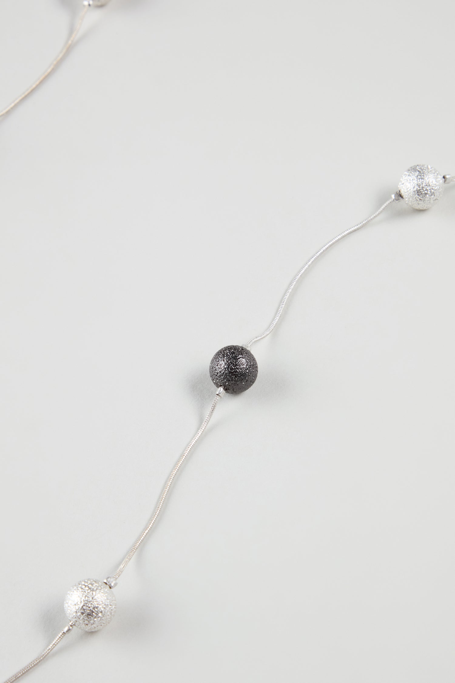 Displayed on a plain light gray background, the Sparkle Spheres Necklace features a slim silver chain adorned with textured silver and black round beads spaced along its length.
