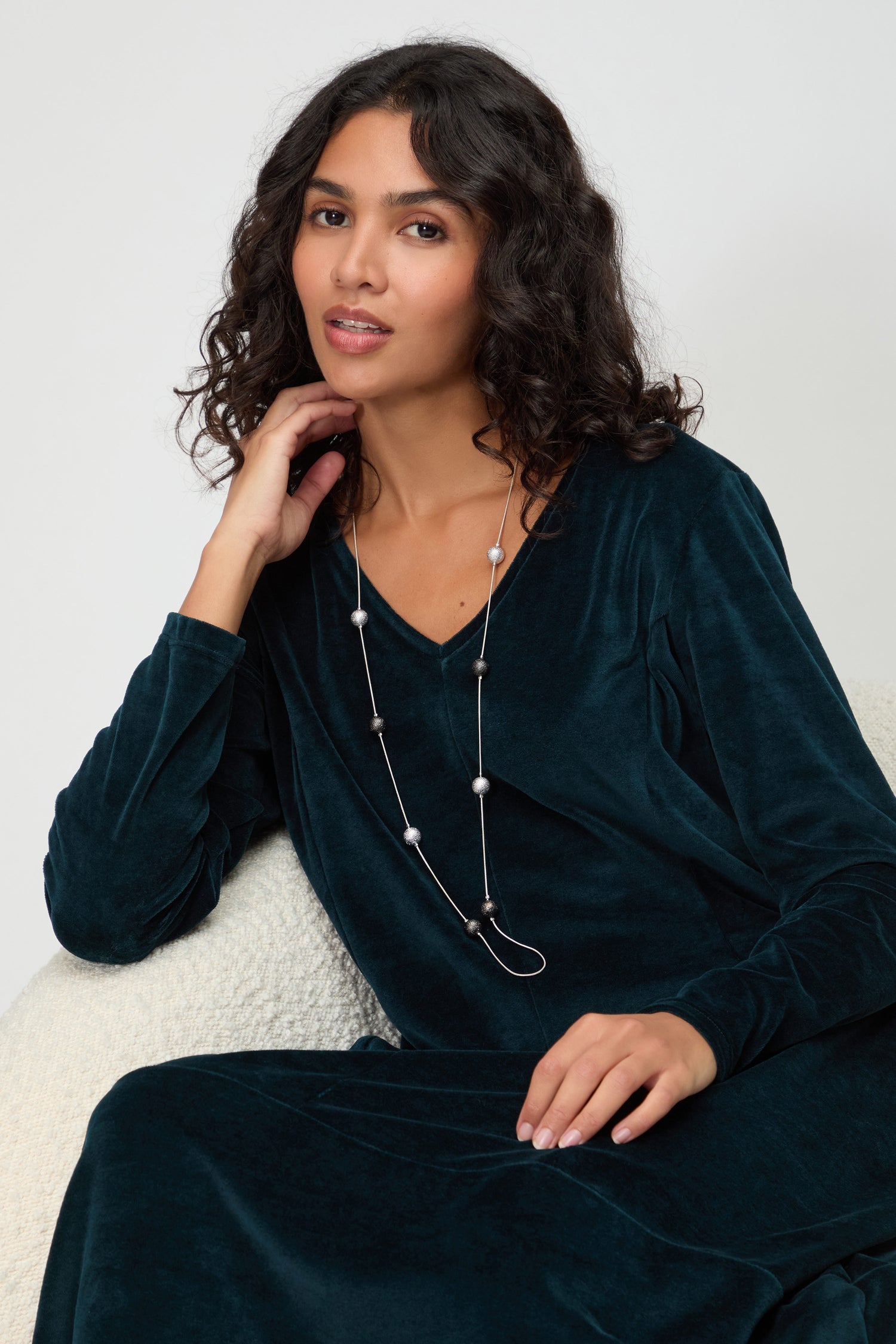 A woman with wavy dark hair in a dark teal velvet dress sits on a light chair, resting her hand on her chin and wearing the Sparkle Spheres Necklace.