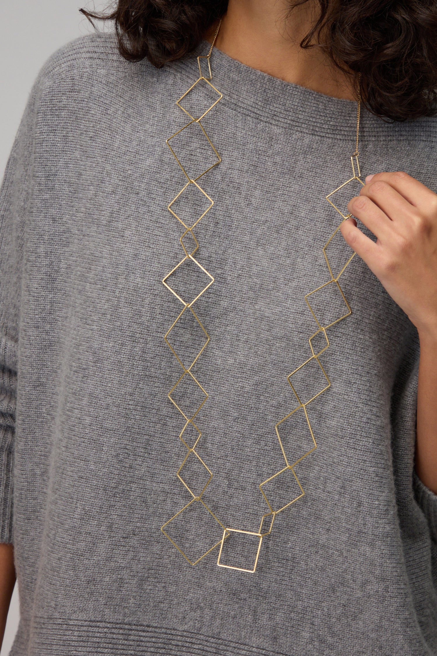 A person wearing a loose grey sweater and the Gold Long Squares Necklace, featuring interconnected gold-toned squares, holding part of the necklace with one hand.