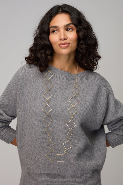Woman with wavy dark hair wearing a loose gray sweater and the Gold Long Squares Necklace, standing with her hands behind her back.