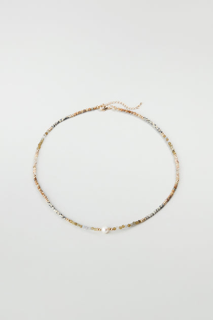 The Short Pearl Bead Necklace features delicate, multicolored semi-precious beads on a thin gold chain with an adjustable clasp, displayed in a circle on a simple light background.