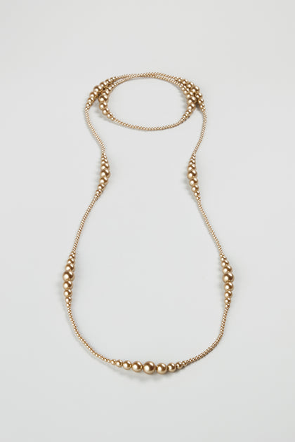 The Graduated Pearl Necklace features small and large round gold-colored beads in a symmetrical pattern, arranged as a long strand and displayed flat on a light background.
