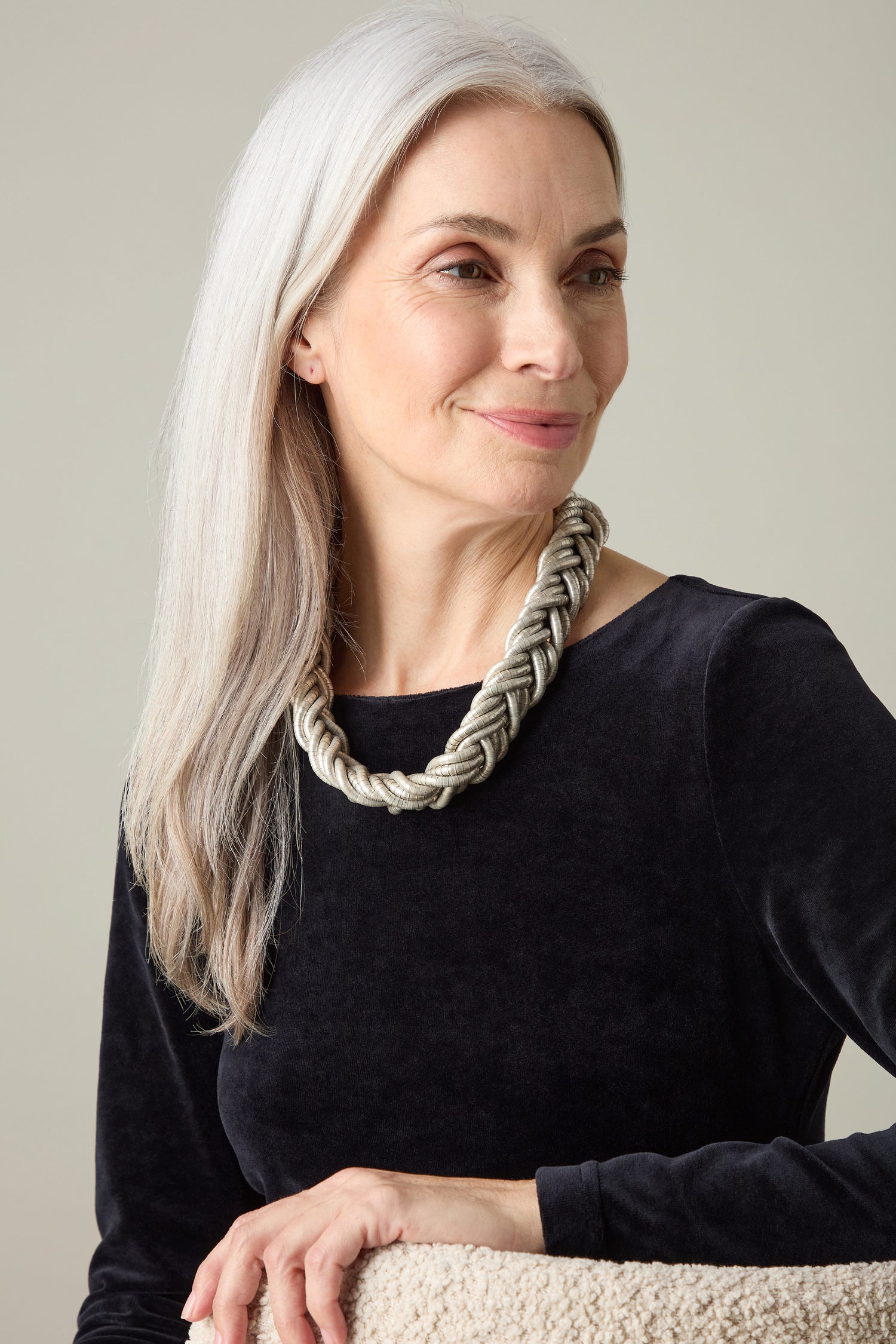 A woman with long grey hair embodies modern elegance as she wears a black top paired with a Silver Braided Necklace, highlighting its artisanal craftsmanship while gazing thoughtfully to the side.