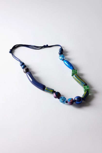 Discover the Painted Shapes Necklace, an artistic statement featuring hand-painted beads in unique shapes and shades of blue, green, and purple, elegantly set against a light gray surface.