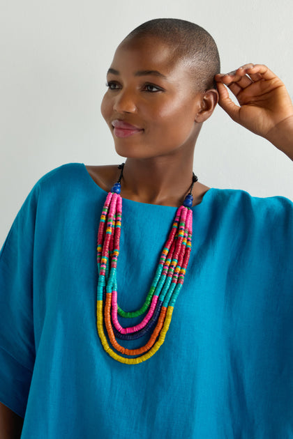 A person in a blue shirt smiles, adjusting the Cascade Rainbow Disc Necklace made of multi-colored disc beads and an adjustable cord closure, against a plain background.