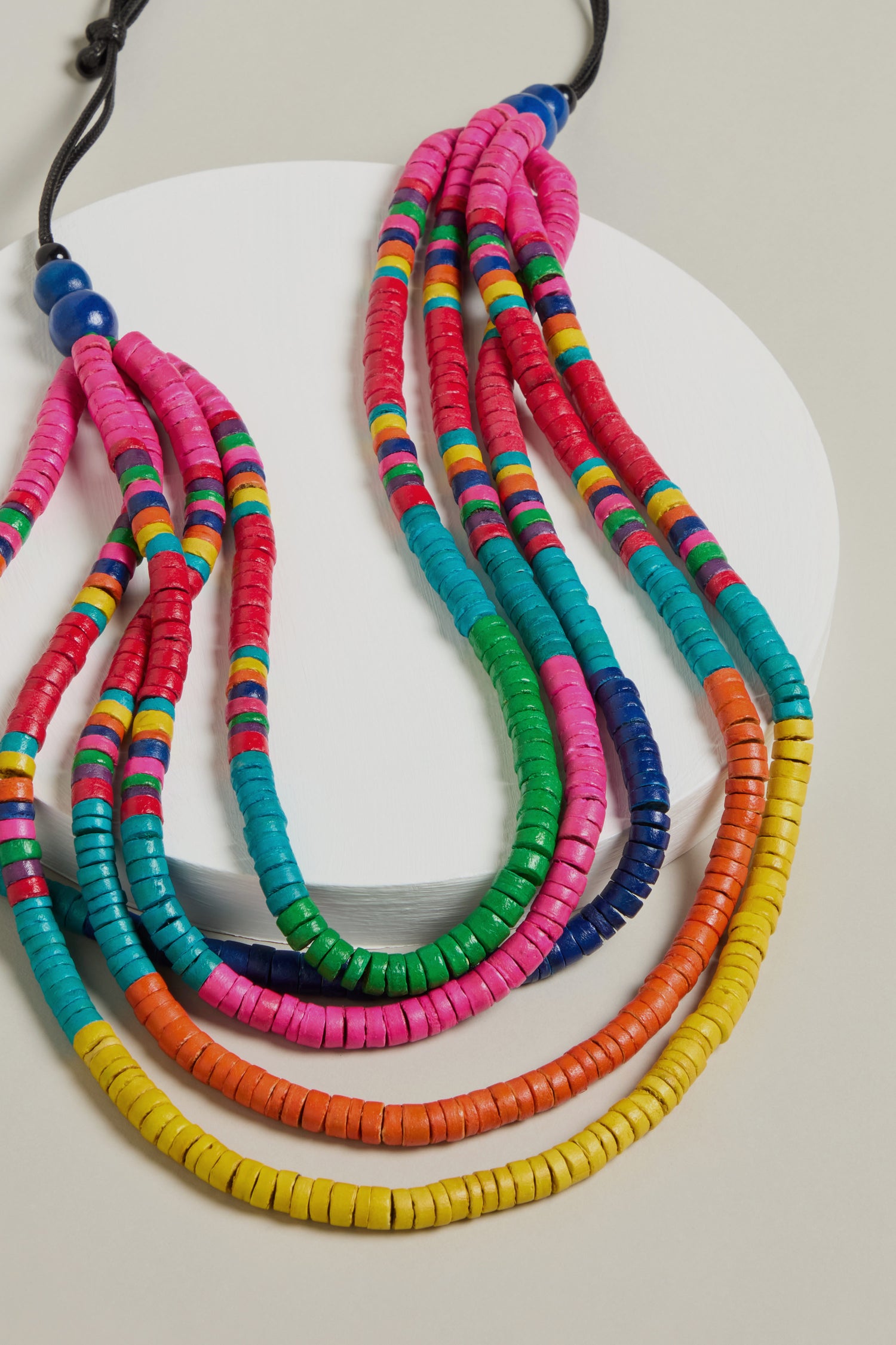 The Cascade Rainbow Disc Necklace, featuring vibrant disc beads in pink, red, blue, green, and yellow tones, elegantly rests on a round white platform. It has an adjustable cord closure for a perfect fit against a neutral background.