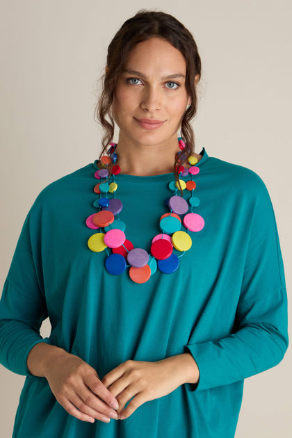 A woman with wavy hair wears a teal top and a vibrant Cascade Rainbow Spheres Necklace adorned with round, flat discs. She is standing against a plain background.