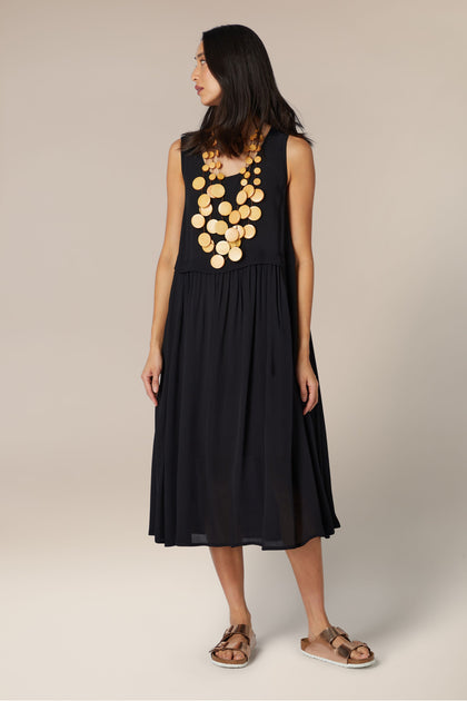 Woman in a sleeveless black midi dress with a pleated skirt and decorative neckline, paired with brown sandals and sporting a Waterfall Spheres Necklace, standing in front of a neutral background.