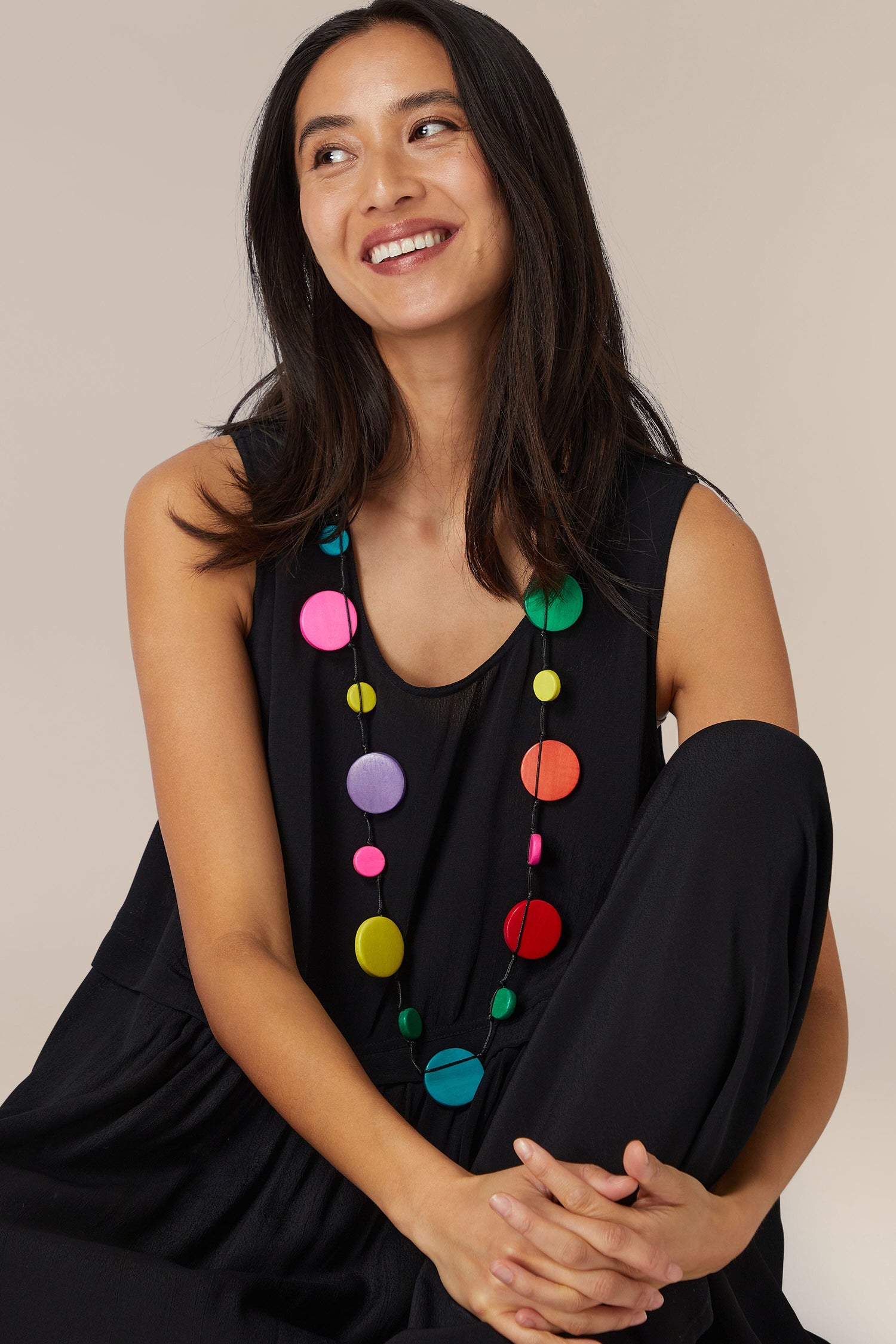 A woman in a black outfit with an adjustable Saturn’s Ring Necklace, featuring wooden discs, smiles while sitting against a neutral background.