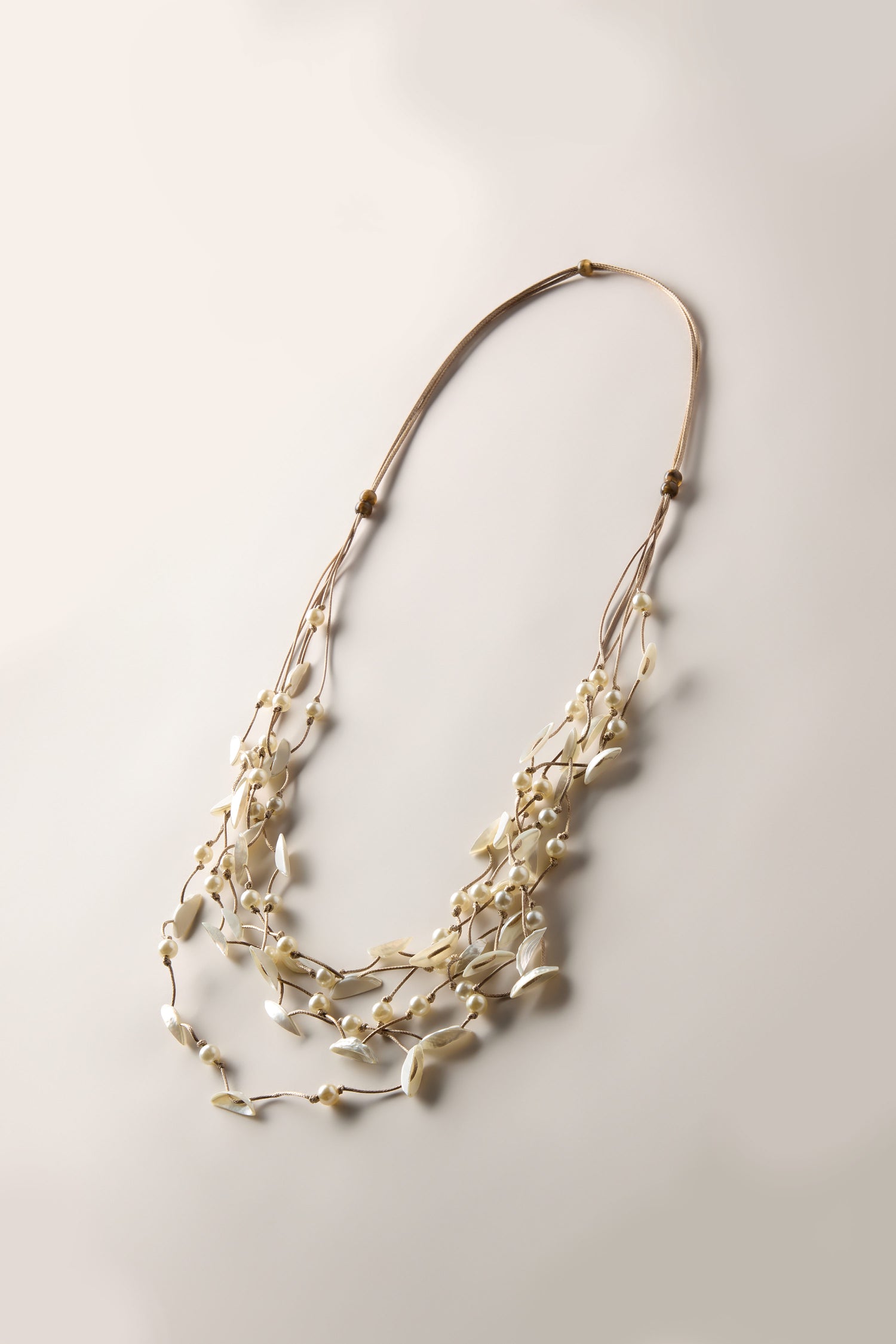 The Cascade Pearl Necklace, a multi-strand contemporary piece featuring white shell beads and a brown cord, is elegantly arranged on a light beige background. This lightweight jewelry piece captures attention with its sophisticated design.