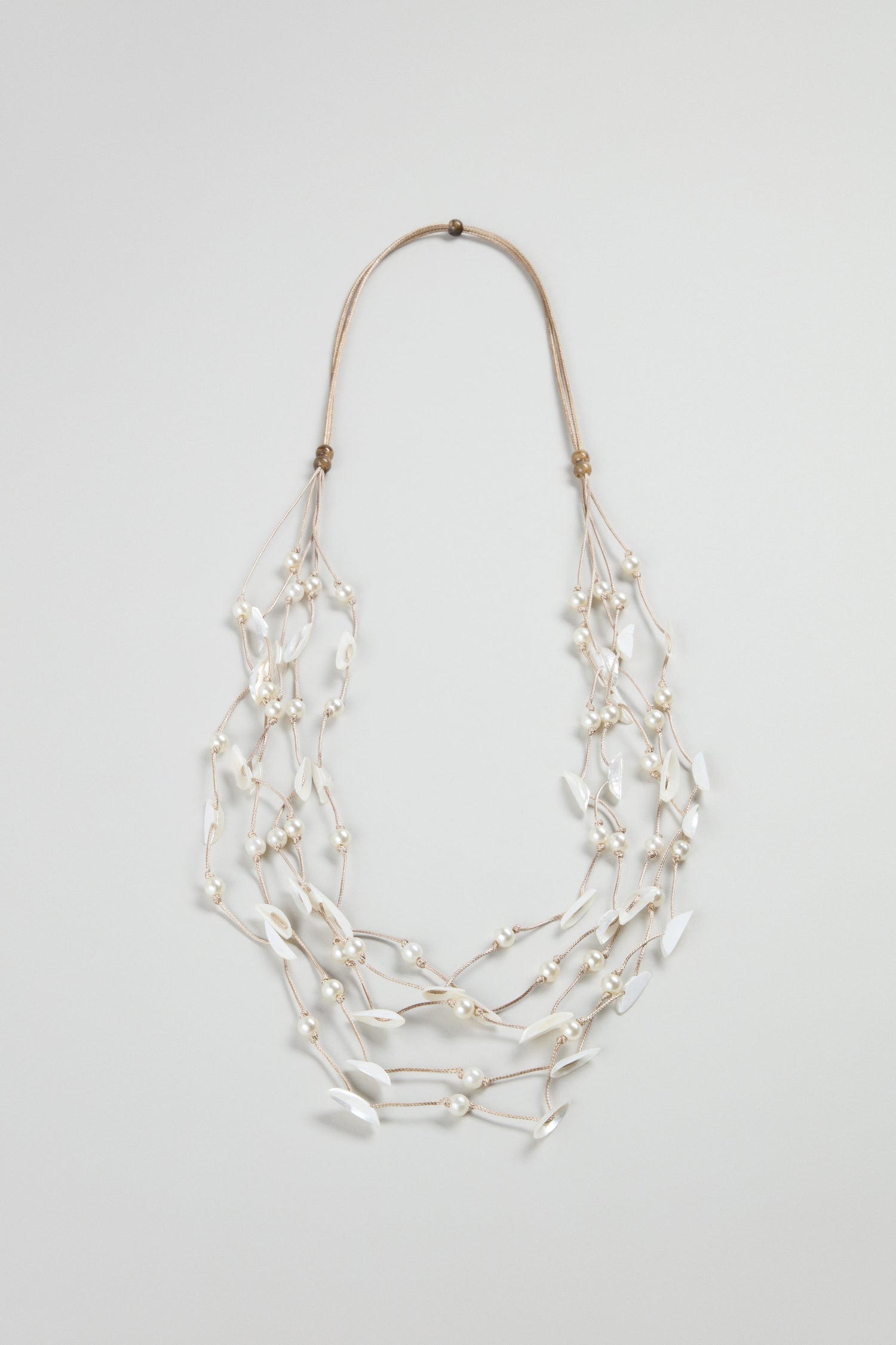 The Cascade Pearl Necklace is a versatile multi-strand piece with white beads and pearl-like leaf shapes, all linked by thin beige cords, beautifully displayed on a plain background.