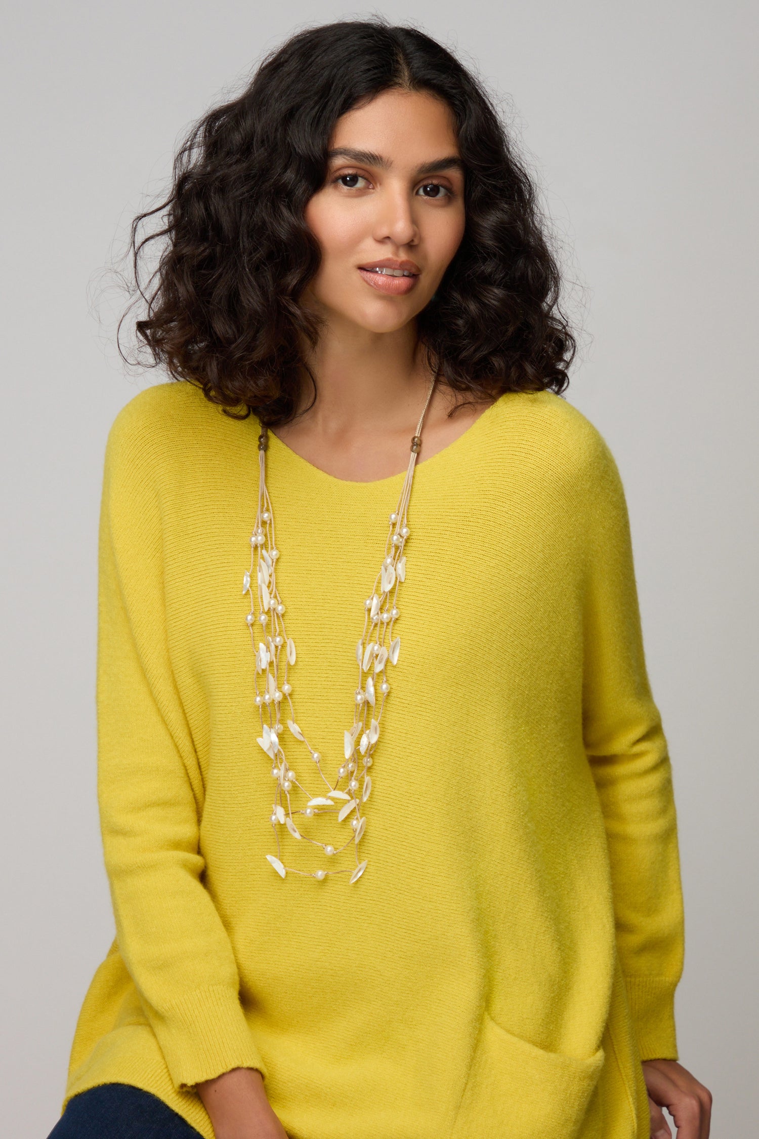 A woman with curly dark hair wears a loose yellow sweater and the Cascade Pearl Necklace, featuring delicate strands, seated against a plain light background.