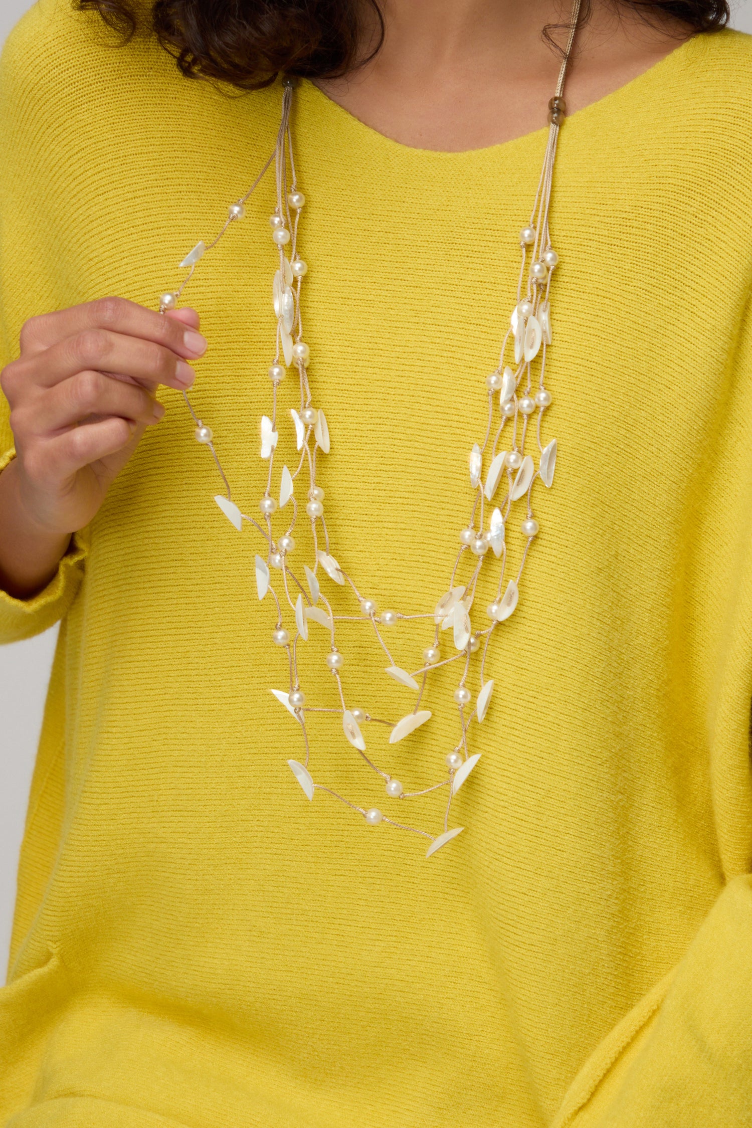 Wearing a yellow sweater, someone holds the Cascade Pearl Necklace, featuring delicate layers of pearl-like beads and leaf-shaped accents that exude timeless radiance.